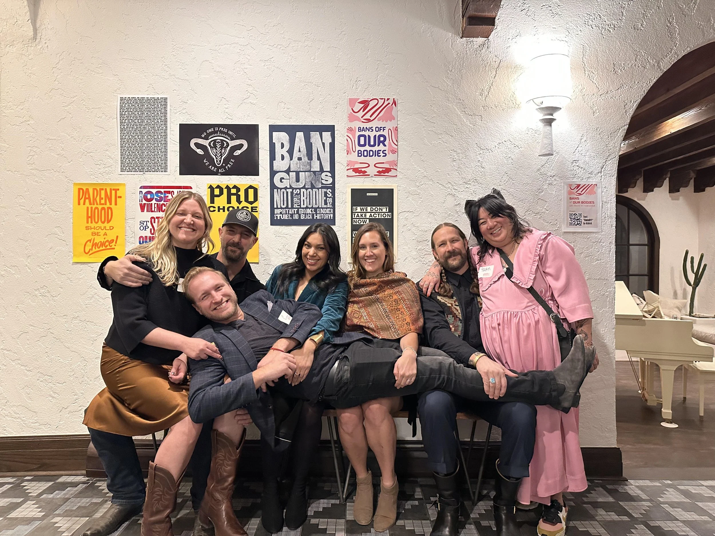 Group of seven diverse people posing together indoors, some sitting and some standing, with posters and protest signs on the wall behind them.