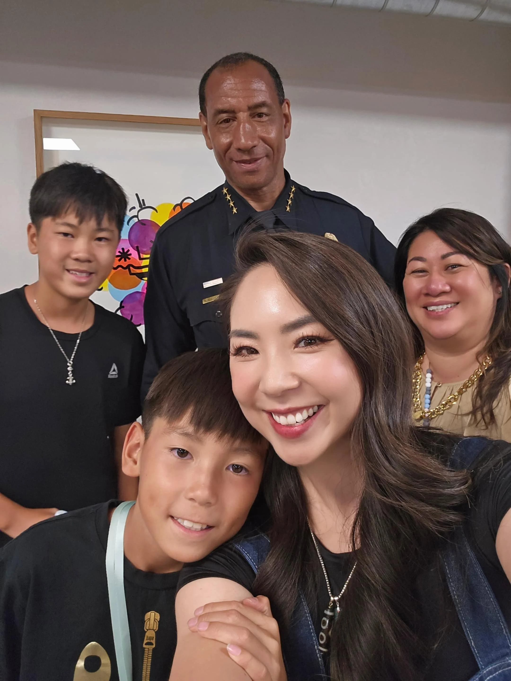 A group selfie featuring six people, including a police officer, young boys, and women, smiling together indoors with a colorful abstract art piece in the background.