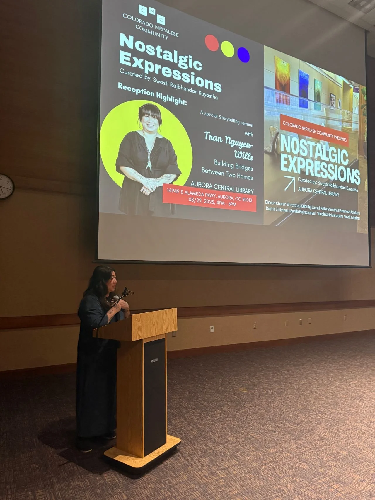 A woman stands at a wooden podium with a microphone, giving a presentation at a large event room. A large screen behind her displays a presentation slide for 'Nostalgic Expressions,' an event organized by Colorado Nepalese Community at Aurora Central