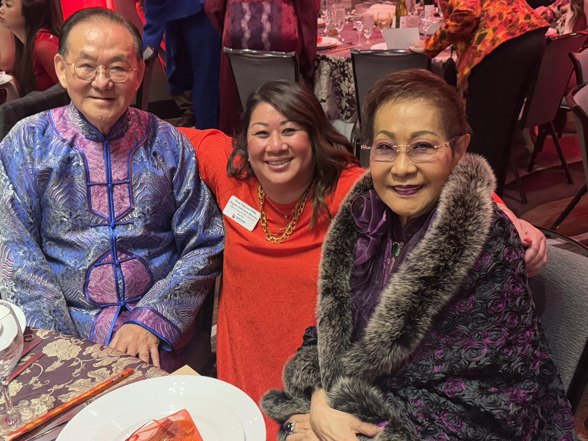 Three people sitting at a banquet table smiling, dressed in colorful formal attire. The man on the left wears a traditional silk jacket, the woman in the middle wears a red dress with gold jewelry, and the woman on the right wears a floral-patterned