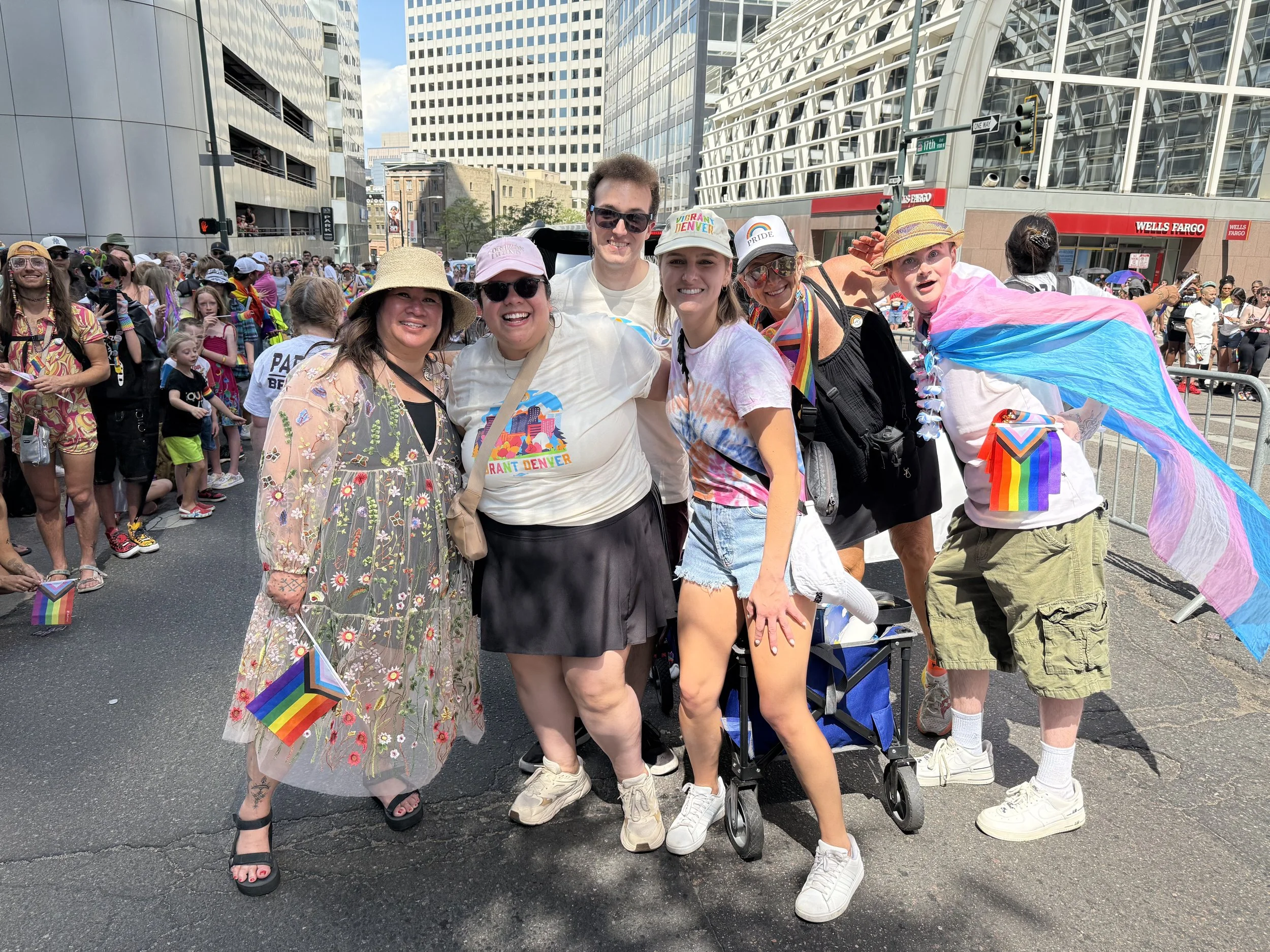 Group of diverse people at a pride parade, wearing casual and festive clothing, holding rainbow flags, with city buildings in the background.