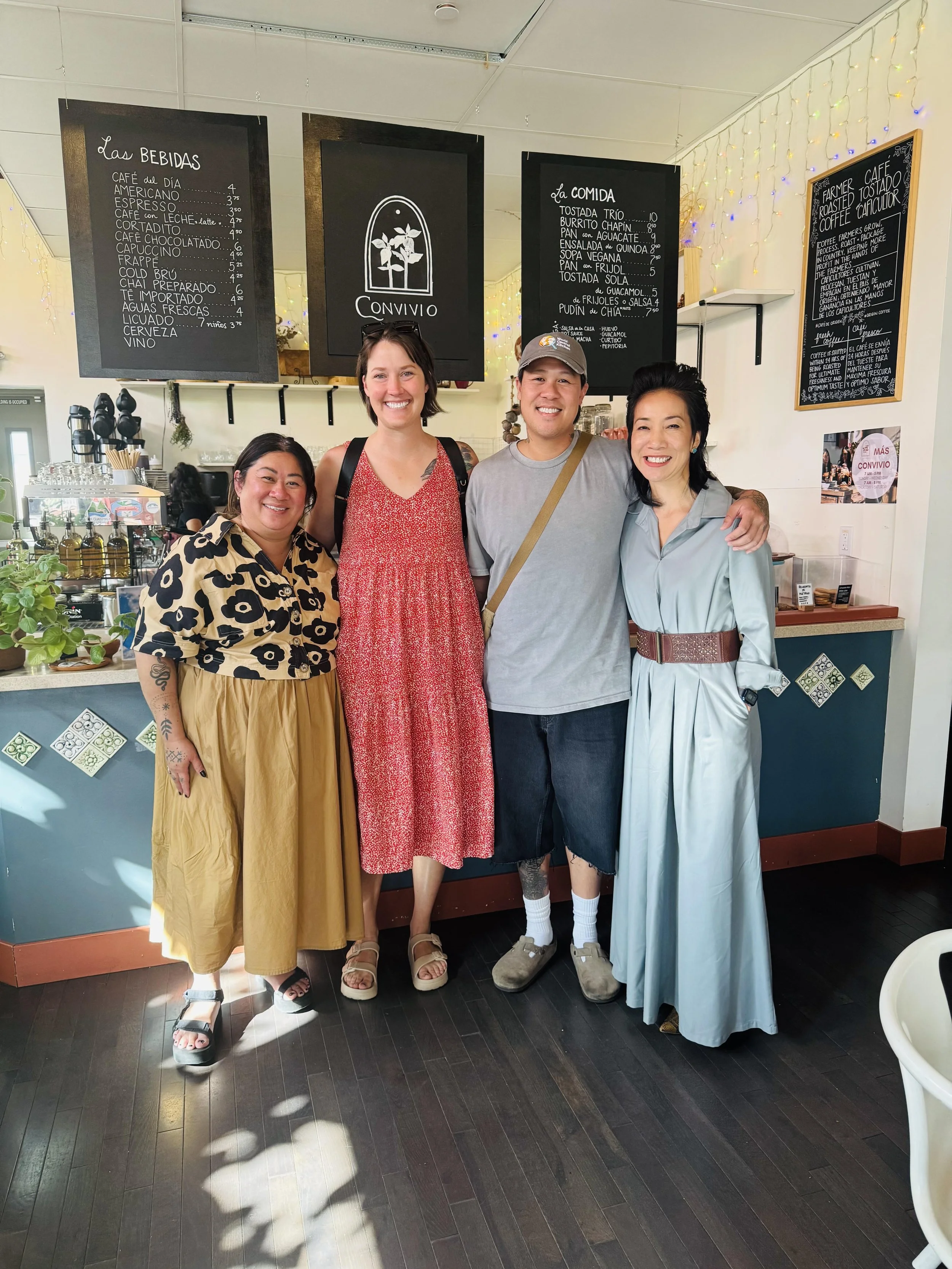 Four people standing together inside a cafe, smiling at the camera, with menu boards on the wall behind them.