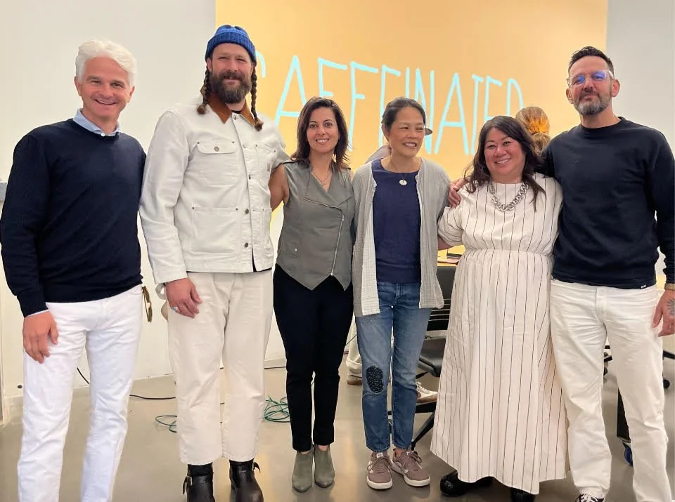 Six diverse people standing close together in an indoor setting with a yellow background that has the word 'AFFINITY' written on it, smiling at the camera.