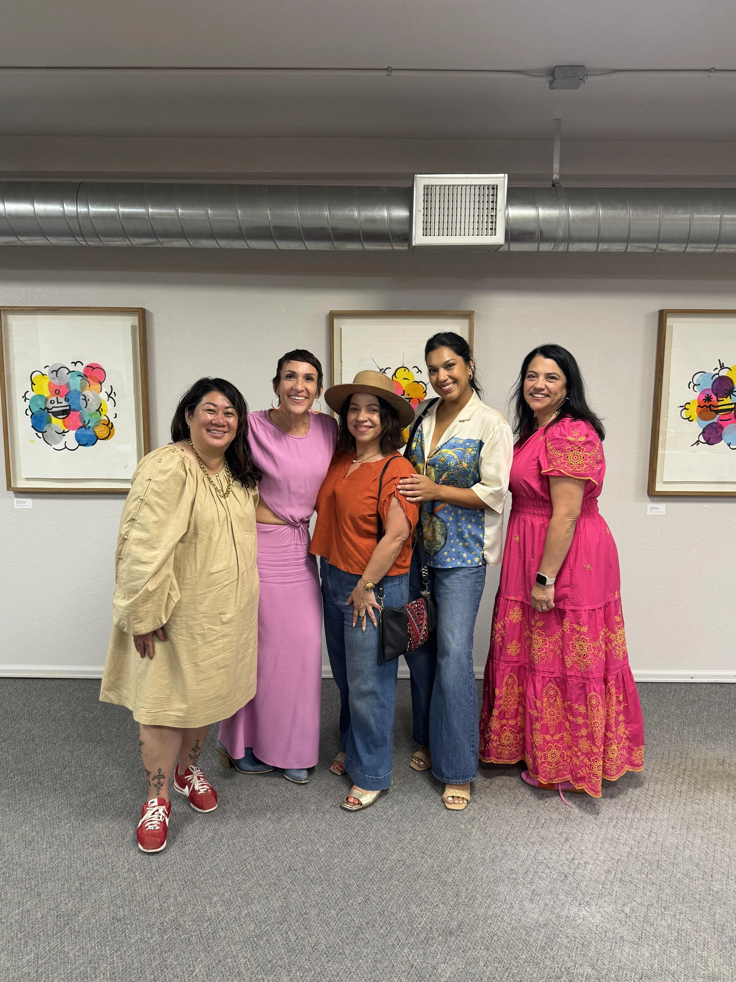 Five women standing together in front of colorful artwork on white walls, smiling at the camera.