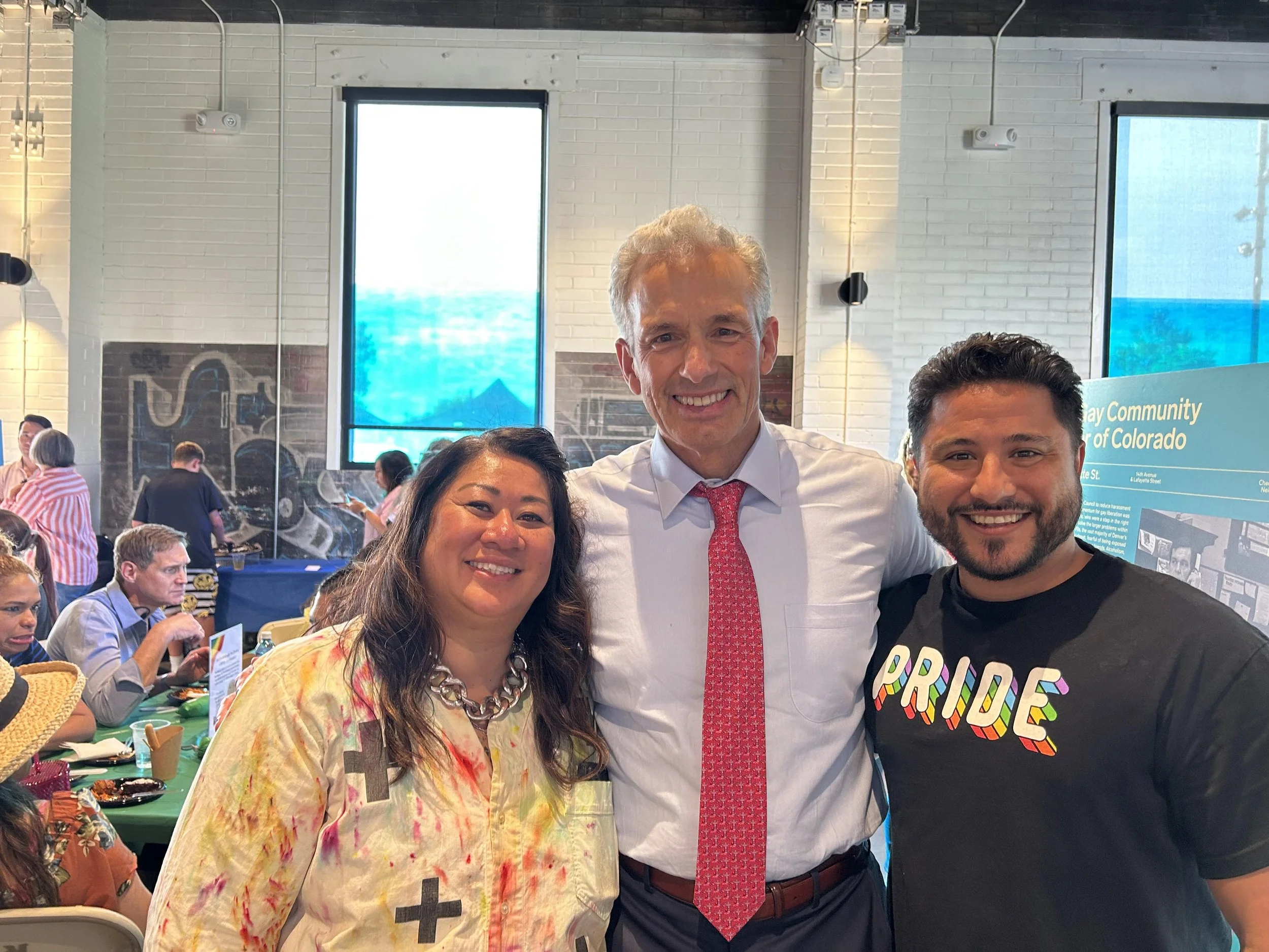 Three smiling people posing at a community event, with a woman on the left wearing a colorful shirt and large chain necklace, a man in the middle with a white shirt and pink tie, and a man on the right wearing a black pride t-shirt. In the background