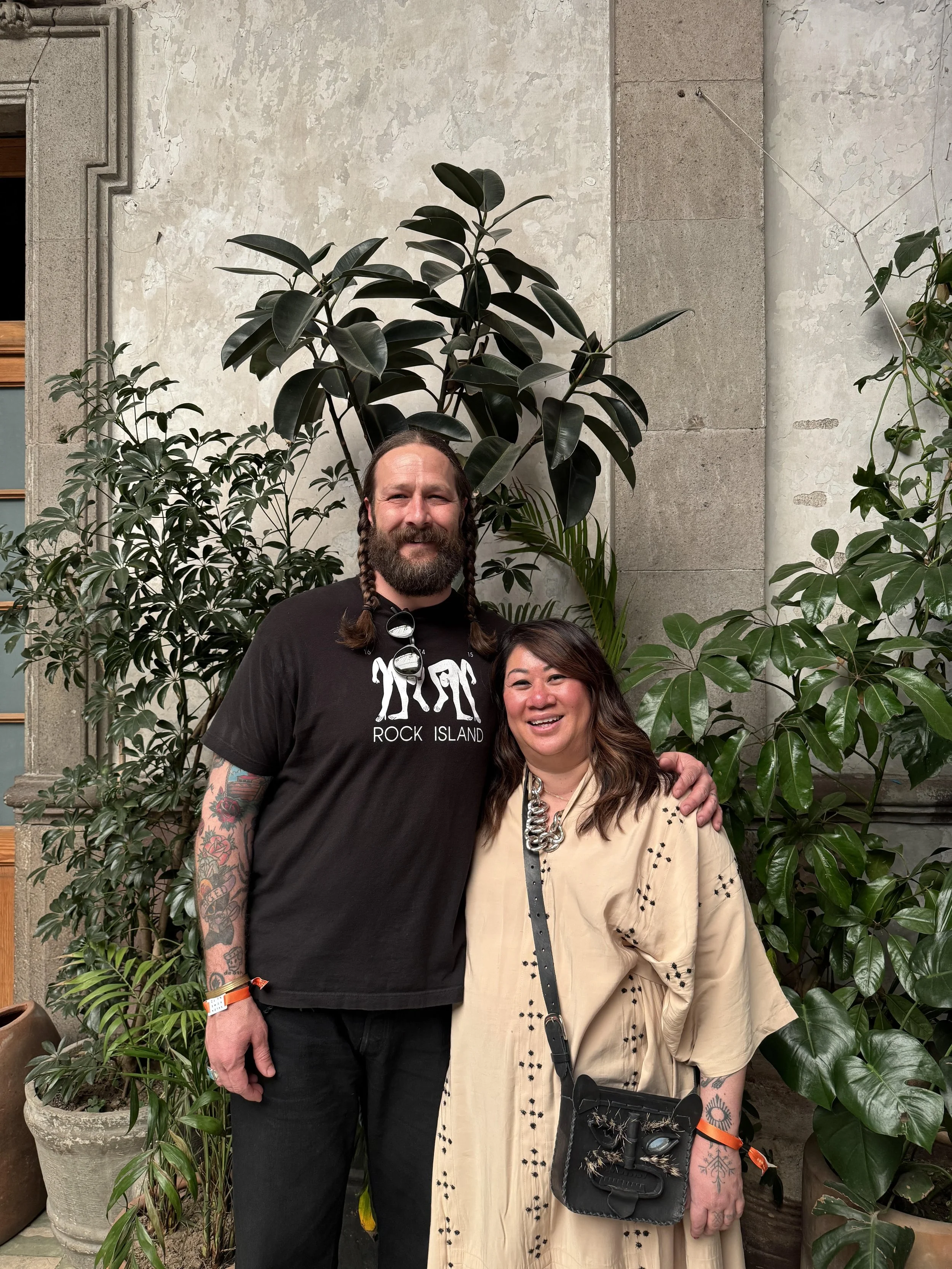 A man and woman standing together in front of large green plants and a stone wall, smiling for the camera.