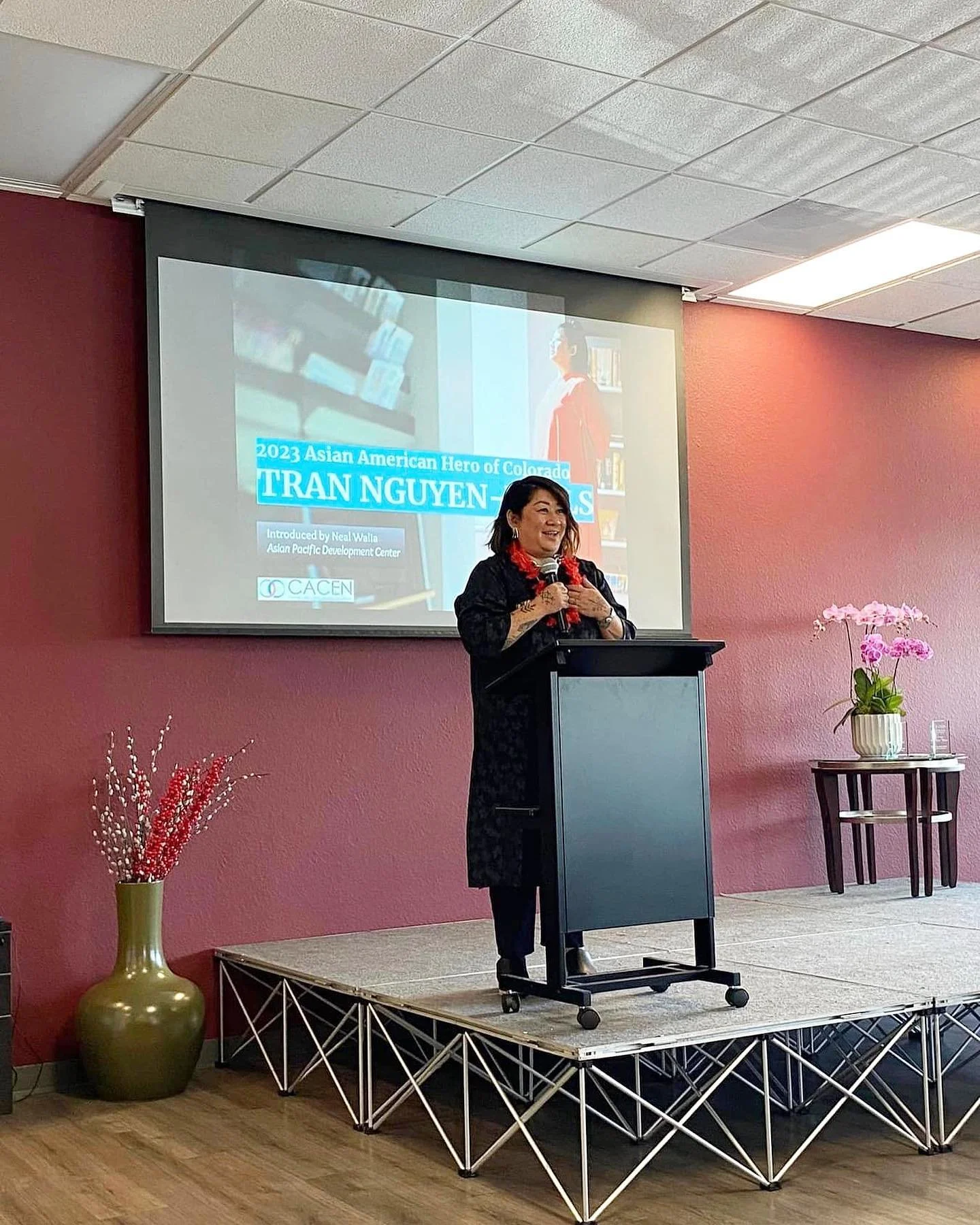 A woman giving a speech at a podium on a small stage, with a projector displaying a slide behind her. The slide features a woman and text about Tran Nguyen-Atkins, a 2023 Asian American Hero of Colorado.
