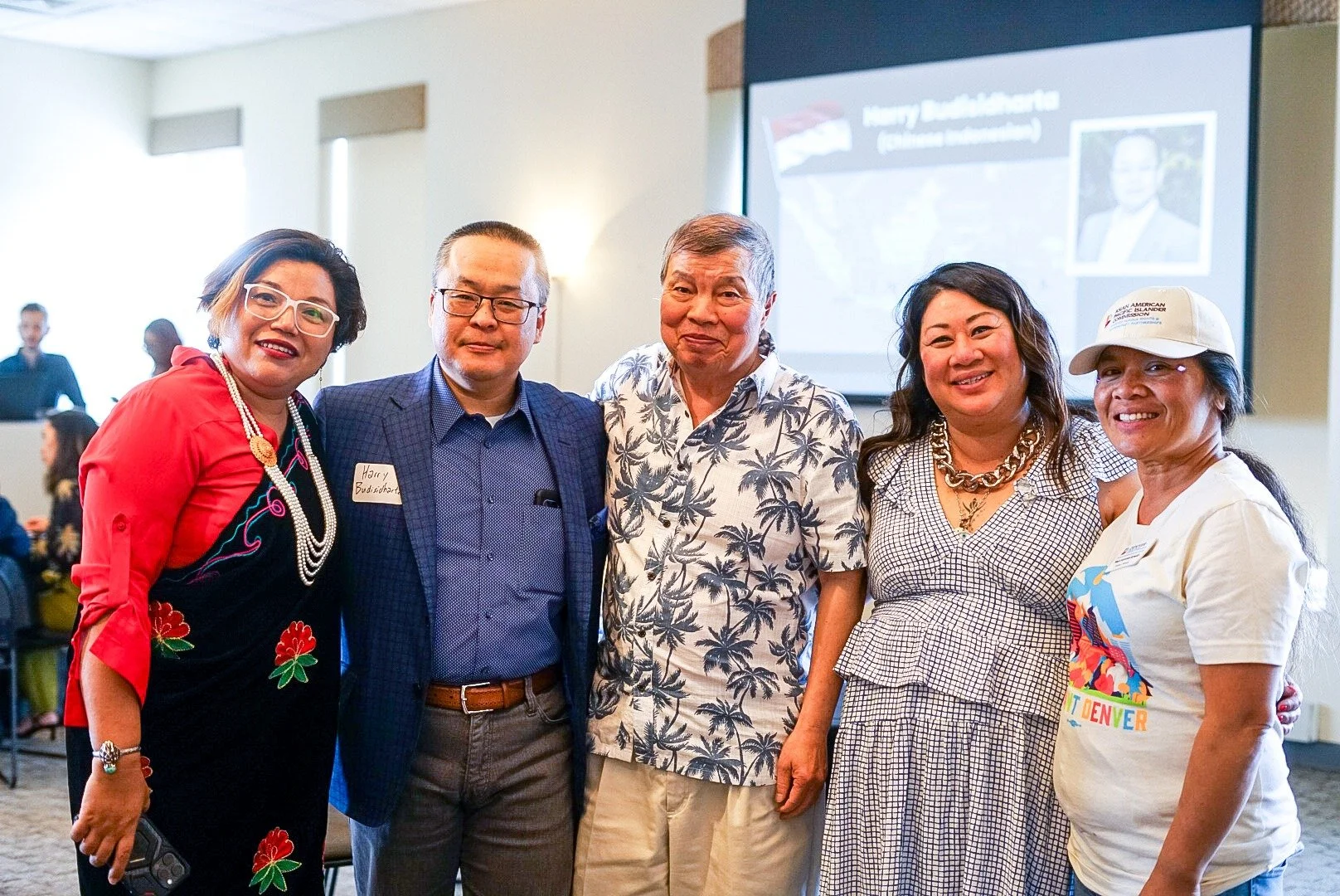 Group of five diverse people smiling and posing for a photo at an indoor event, with a presentation screen in the background.