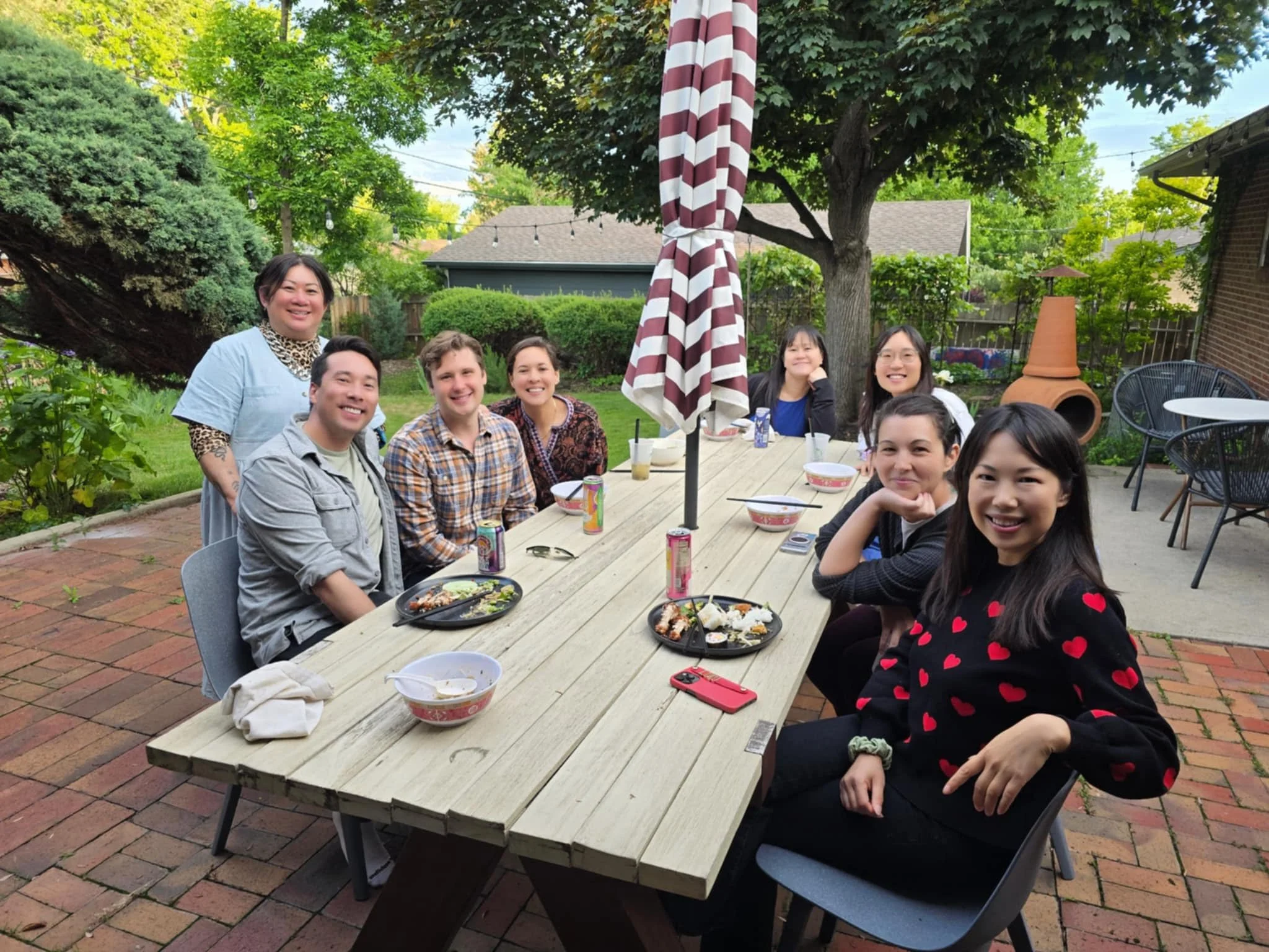 Group of people enjoying a meal outdoors around a long wooden table in a backyard garden with green trees and bushes, some with drinks, food, and cell phones on the table.