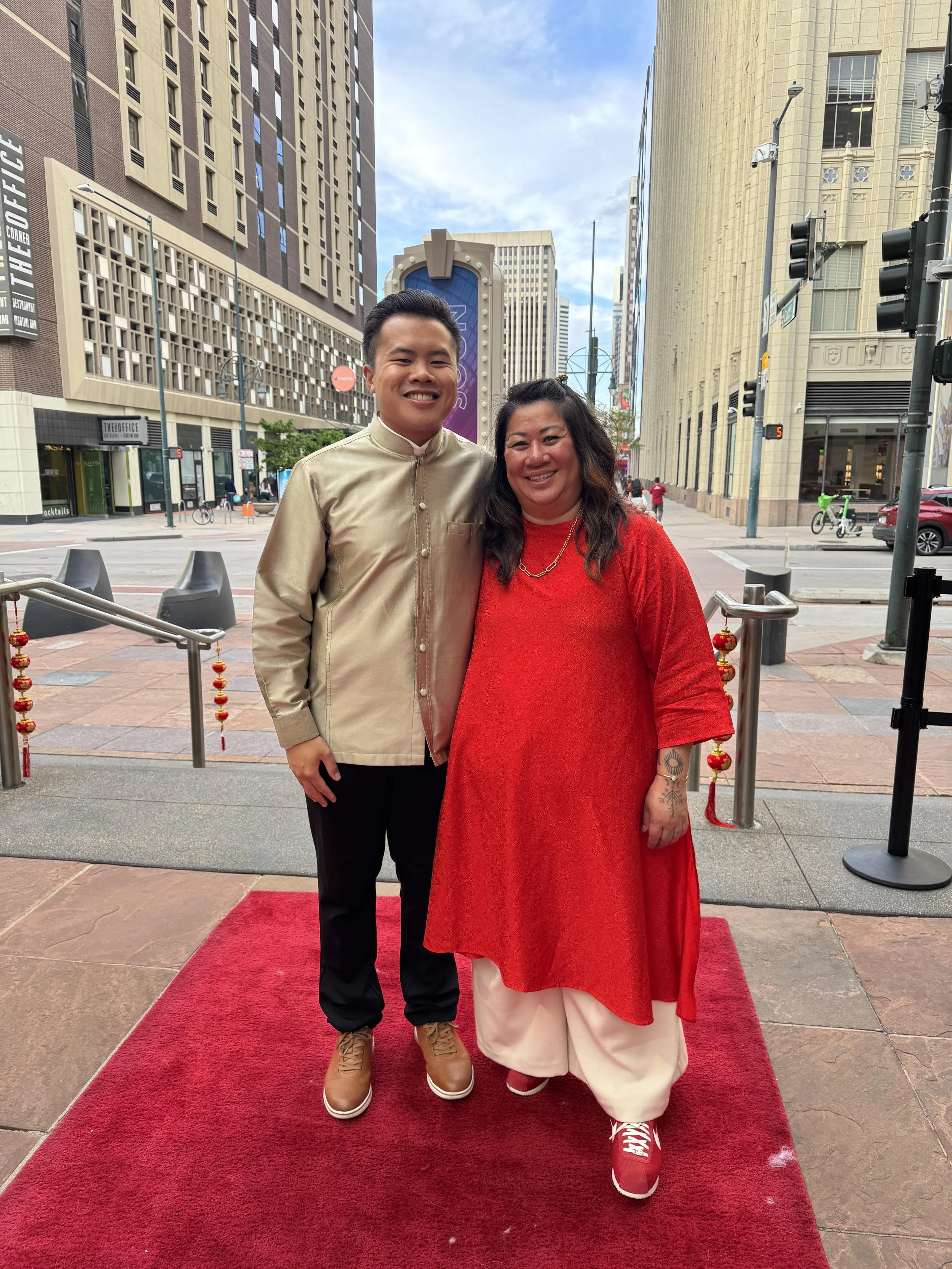 A young man and a woman standing together on a red carpet outside in an urban area, smiling at the camera. The man is wearing a traditional beige outfit, and the woman is dressed in a red top and cream-colored pants, with some tattoos visible on her