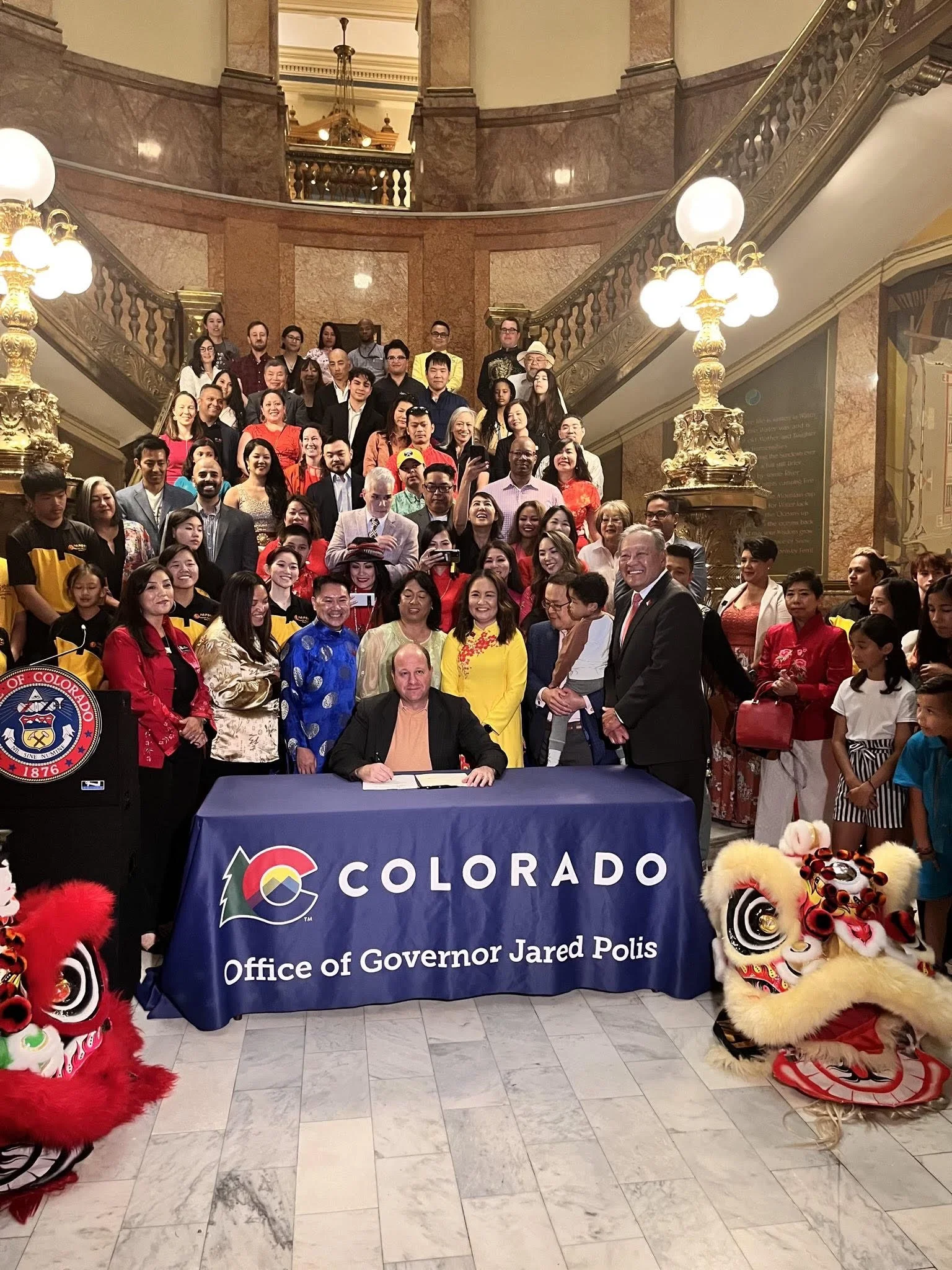 A large group of people gathered in an elegant interior with a staircase and ornate lighting, posing for a photo behind a table with a Colorado-themed banner. The banner reads 'Office of Governor Jared Polis'. Two lion dance costumes are seen on the