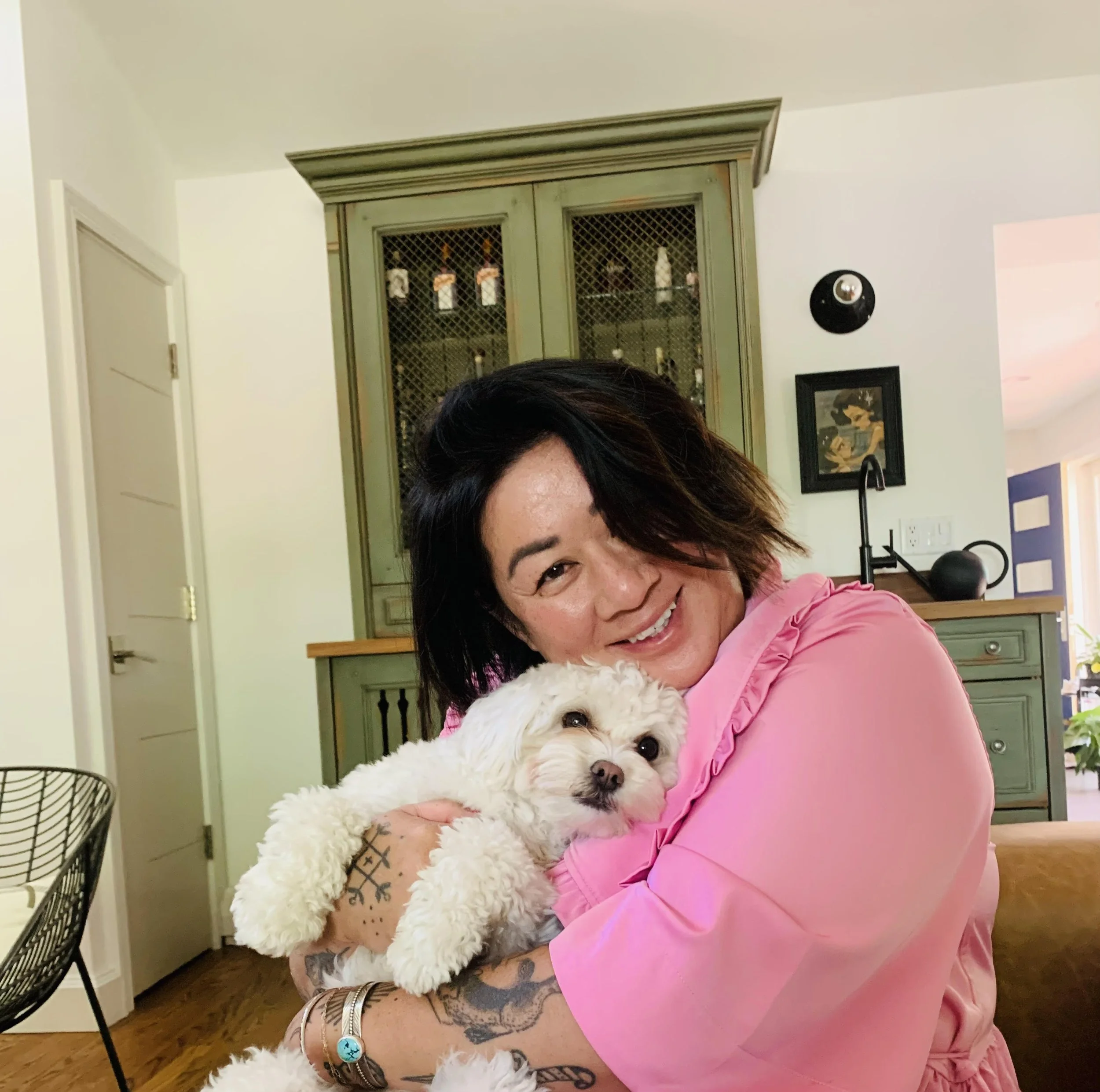 A woman with black hair wearing a pink top is smiling and hugging a small white fluffy dog in a cozy kitchen.