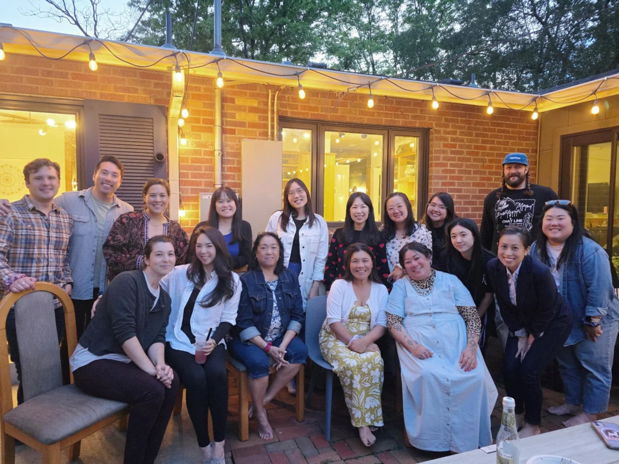 Group of diverse people gathered outdoors on a patio during the evening, smiling for a photo, with string lights hanging overhead and a brick building in the background.