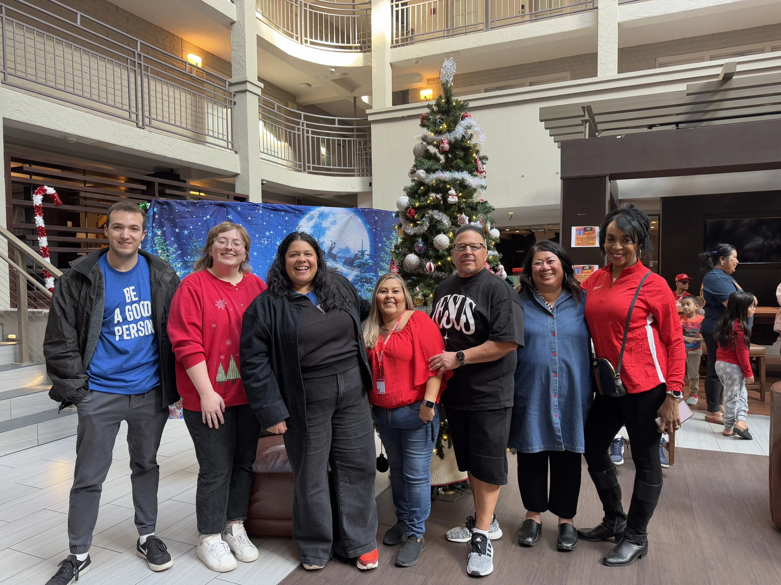 Group of seven people smiling and posing in front of a decorated Christmas tree inside a hotel lobby during the holidays.