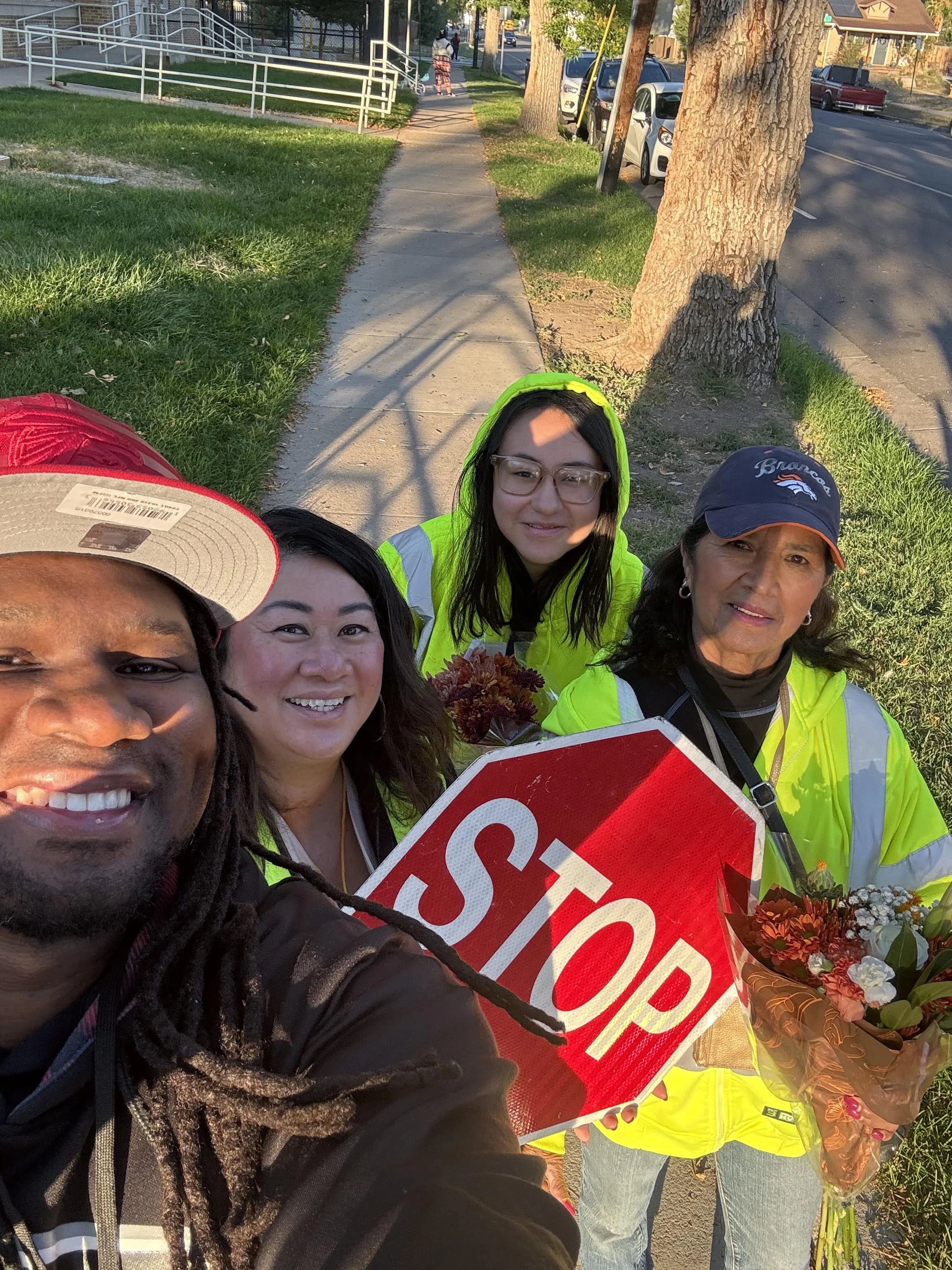 Group of four people taking a selfie on a sidewalk, holding a stop sign and flowers, with trees and parked cars in the background.