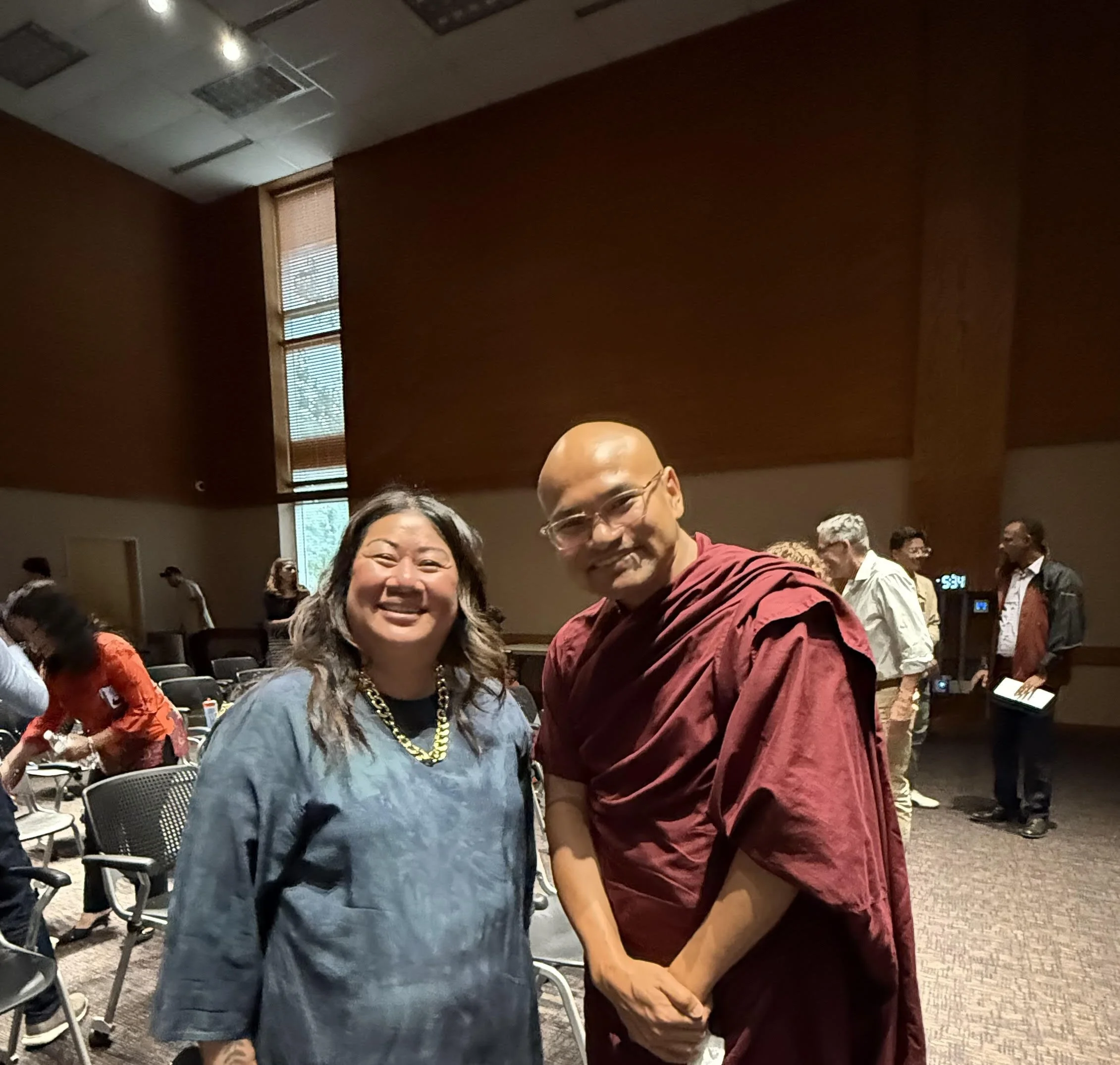 A woman and a man in traditional Buddhist robes smiling at the camera in a large hall or auditorium with other people in the background.