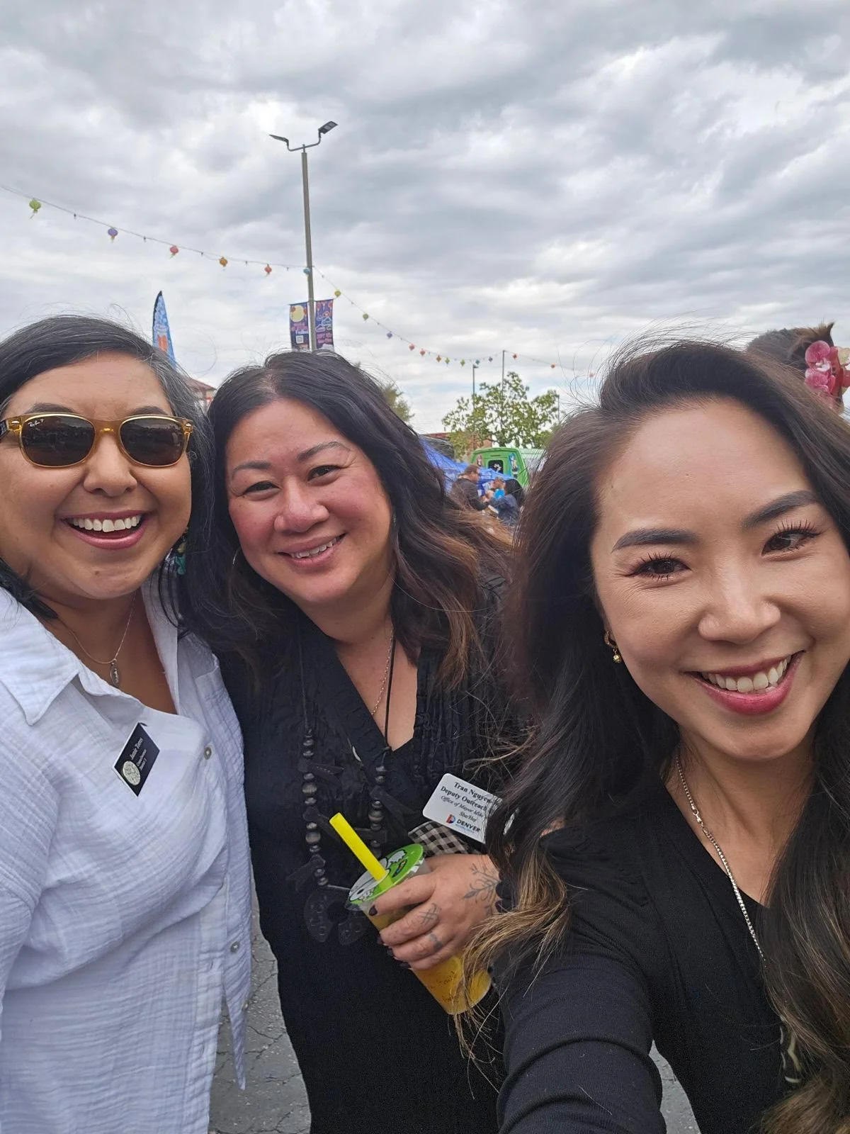 Three women smiling and taking a selfie outdoors at a festival, with cloudy sky and colorful decorations in the background.