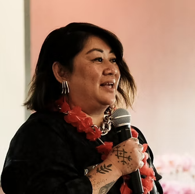 A woman is speaking into a microphone during an indoor event, wearing a black outfit, red floral necklace, and hoop earrings, with a pinkish background.