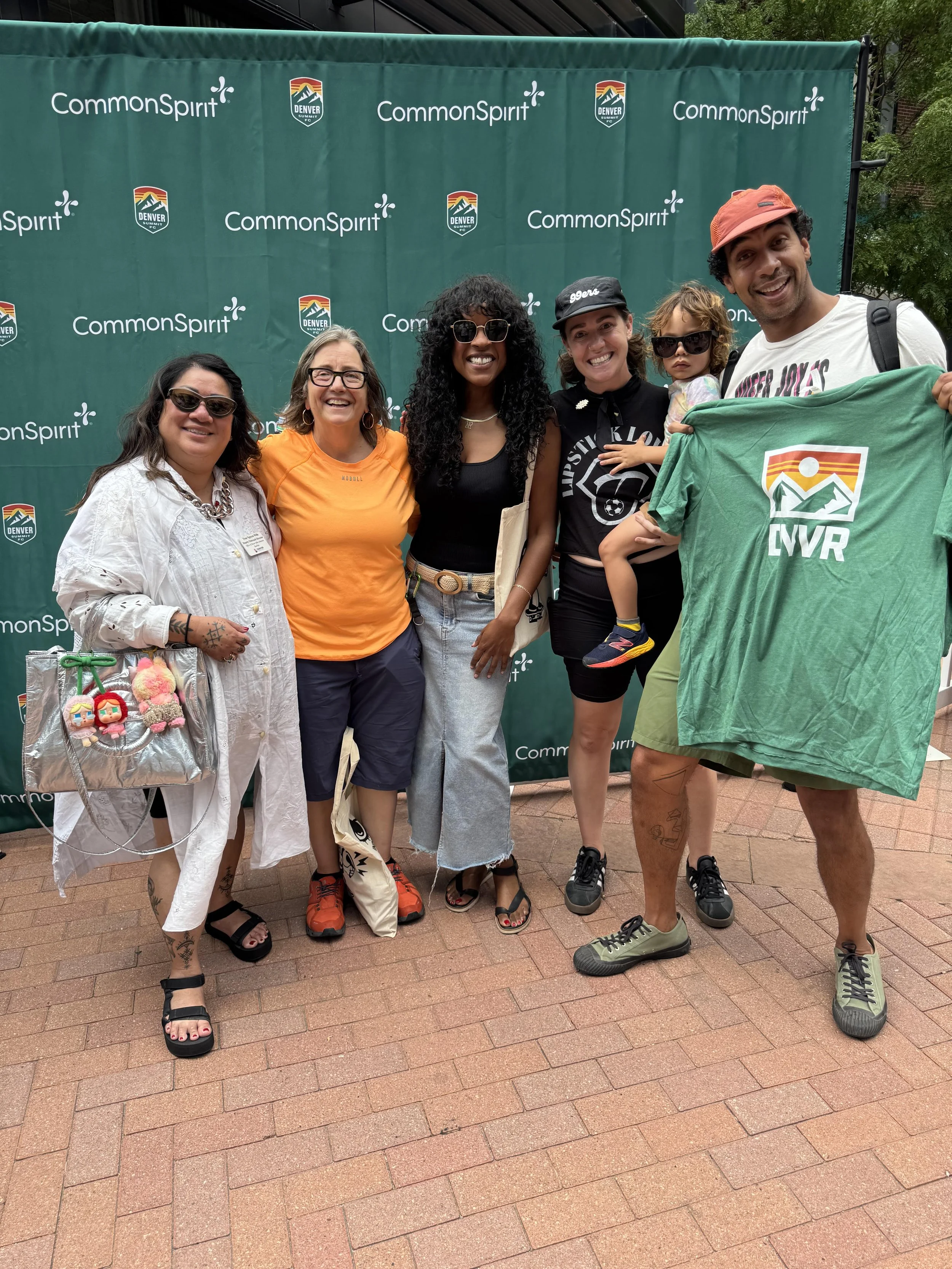 A group of six diverse people, including one child, standing outdoors in front of a green CommonSpirit backdrop, smiling for a photo. They are dressed casually, with some wearing sunglasses and hats, and one person holding a green t-shirt with a colo