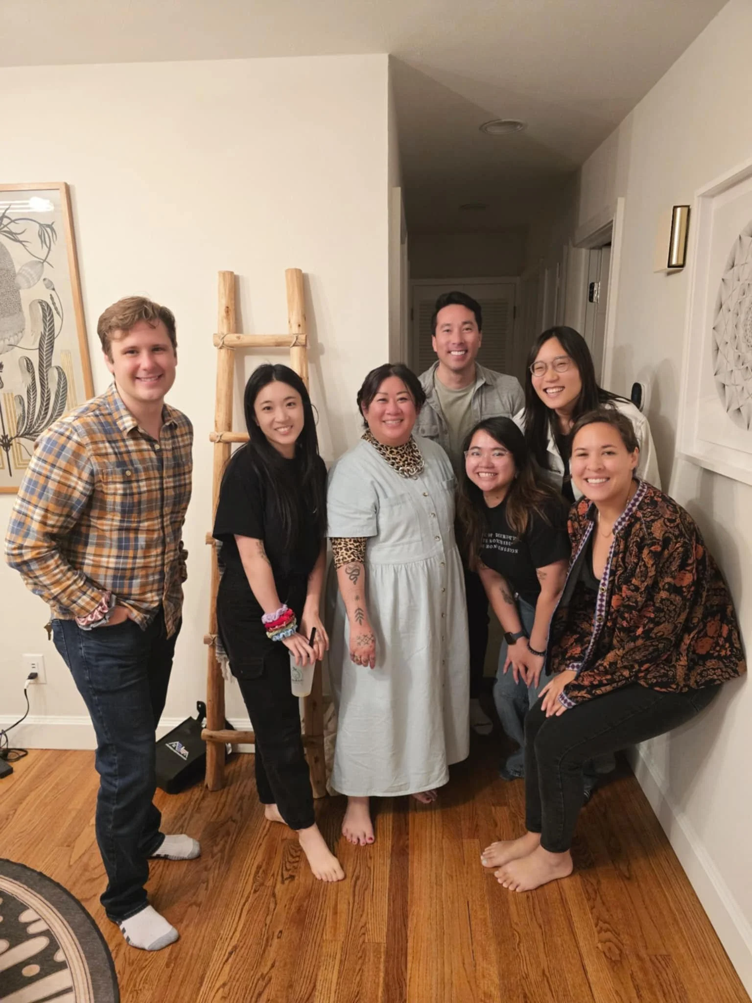 A group of seven people smiling and posing together in a home interior near a wooden ladder and wall art.