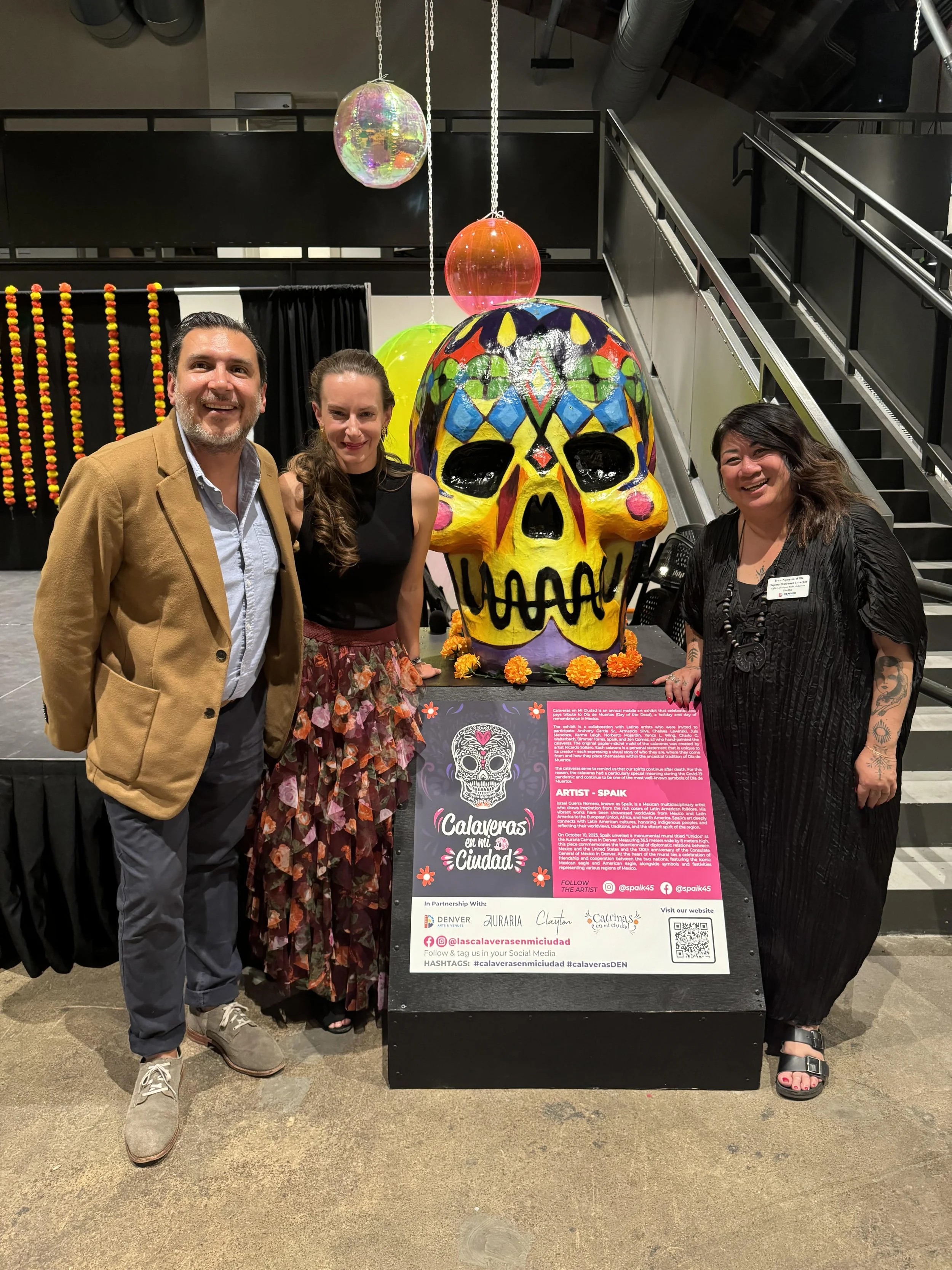 Three people standing next to a colorful decorated skull sculpture at an event celebrating Day of the Dead. The sculpture is placed on a black platform with a descriptive sign in front.