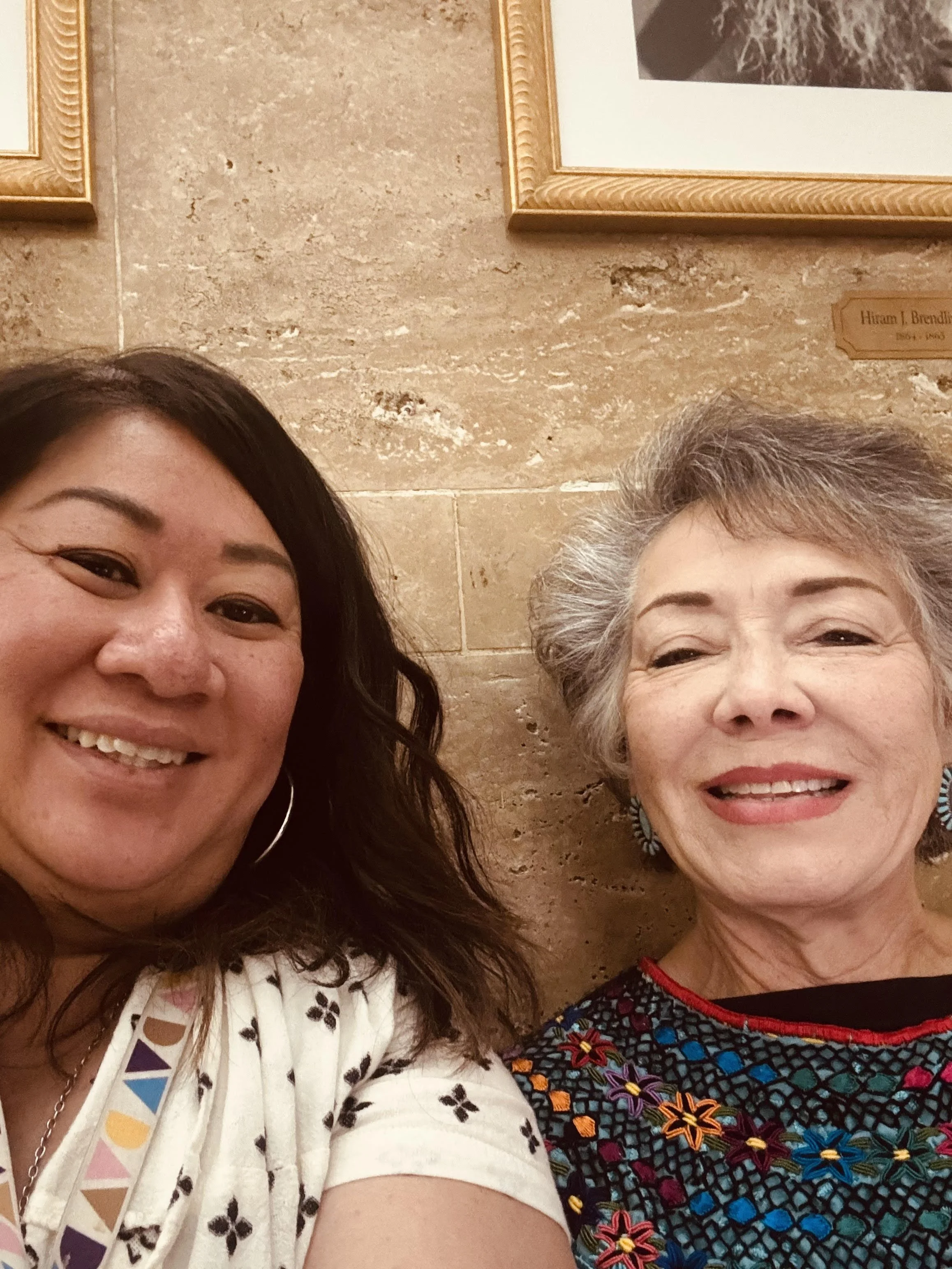 Two women smiling for a selfie, standing in front of a beige stone wall with framed pictures.