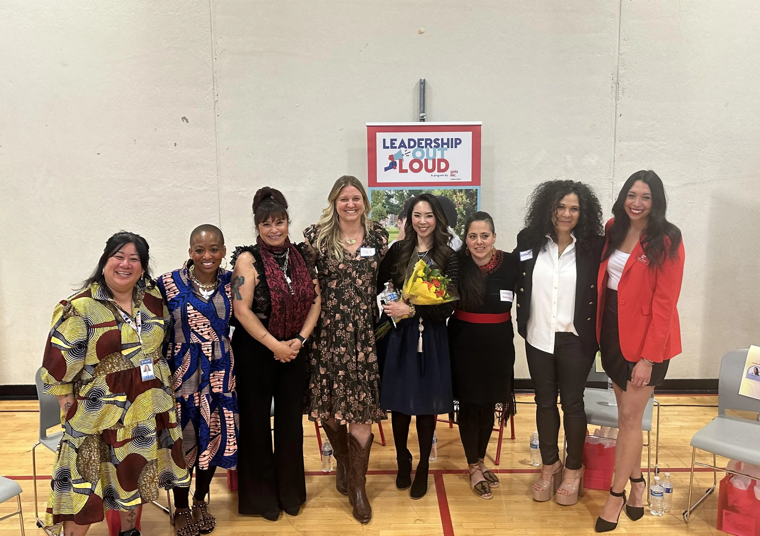 A diverse group of nine women standing together in a gymnasium, smiling, with a sign that reads 'Leadership Out Loud' behind them. One woman is holding a bouquet of flowers.