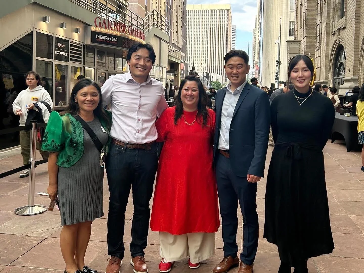 A group of six people standing outside in an urban area, smiling at the camera. They are dressed in casual and semi-formal clothing, with tall buildings and a theatre marquee in the background.