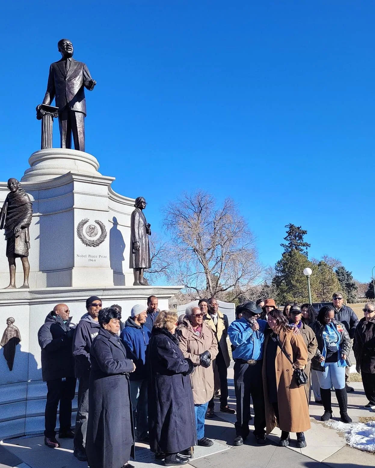 A group of people gathered around a monument featuring statues of prominent historical figures, including Dr. Martin Luther King Jr., with trees and a clear blue sky in the background.