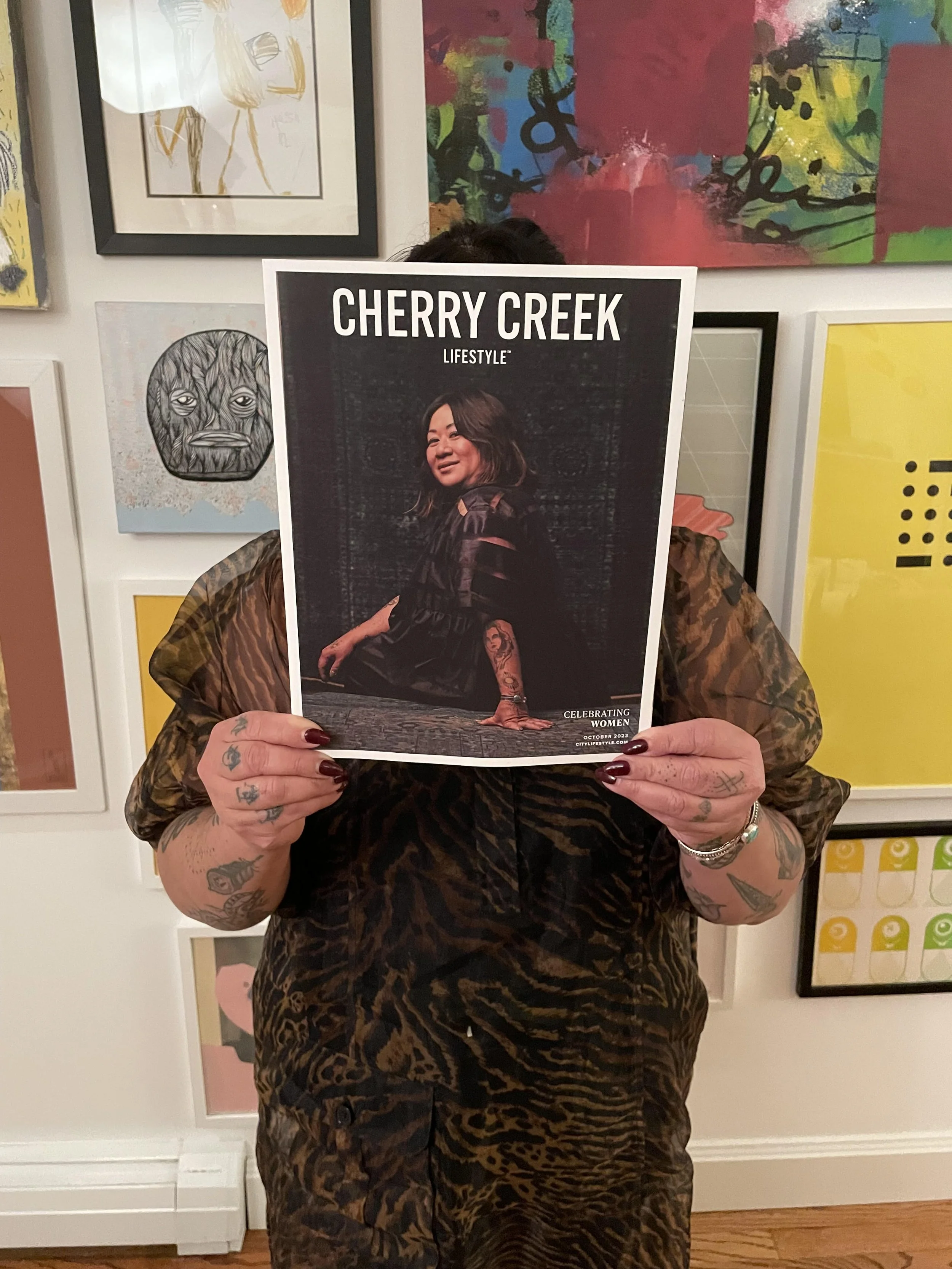 Person holding a magazine titled 'Cherry Creek Lifestyle' in front of face, with tattoos visible on hands and arms, colorful artwork on the wall in the background.
