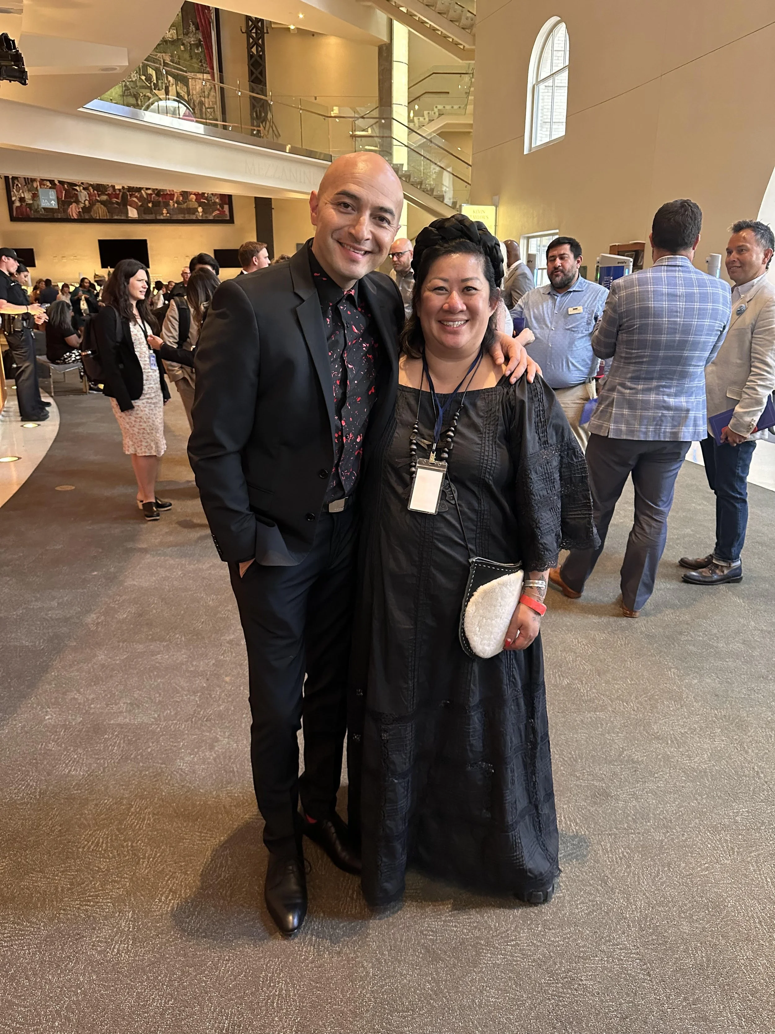 A man and woman smiling at a crowded indoor event, with the man dressed in a black suit and the woman in a black dress holding a white purse, standing together in front of a busy background with people talking and walking.