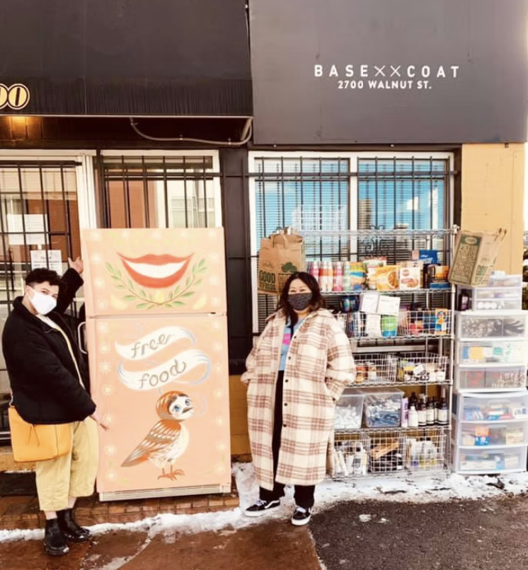 Two women standing outside a free food distribution stand with shelves of food and supplies. They are wearing masks. One woman points at a sign with a cartoon bird and a smiling mouth, indicating free food.