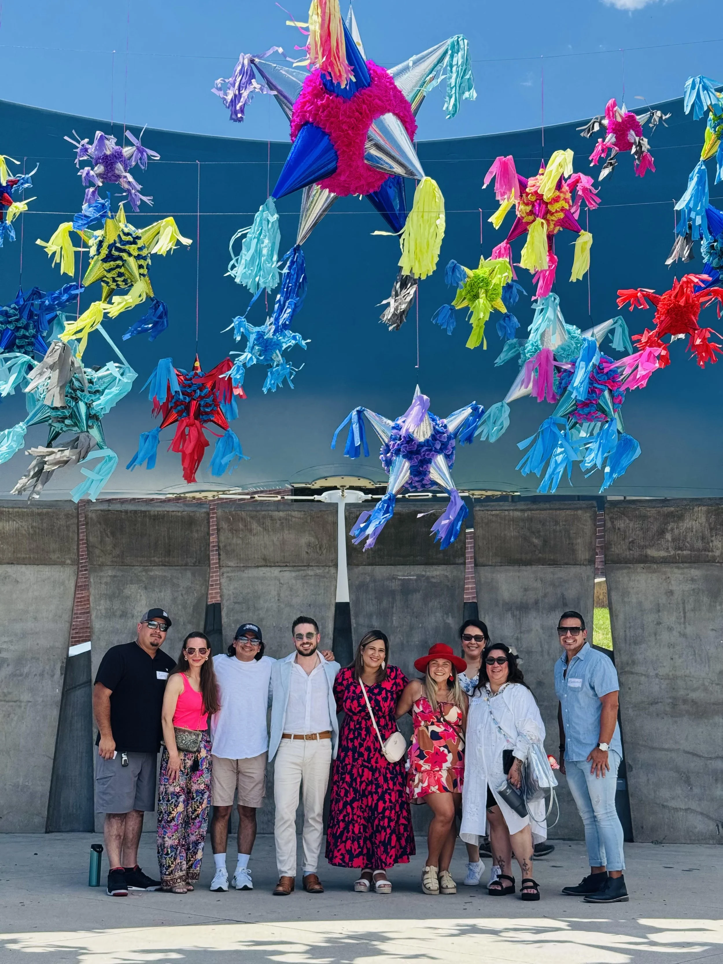 A group of eight people standing in front of a concrete wall, posing for a photo. Above them are colorful, star-shaped piñatas hanging from the ceiling, set against a bright blue sky.