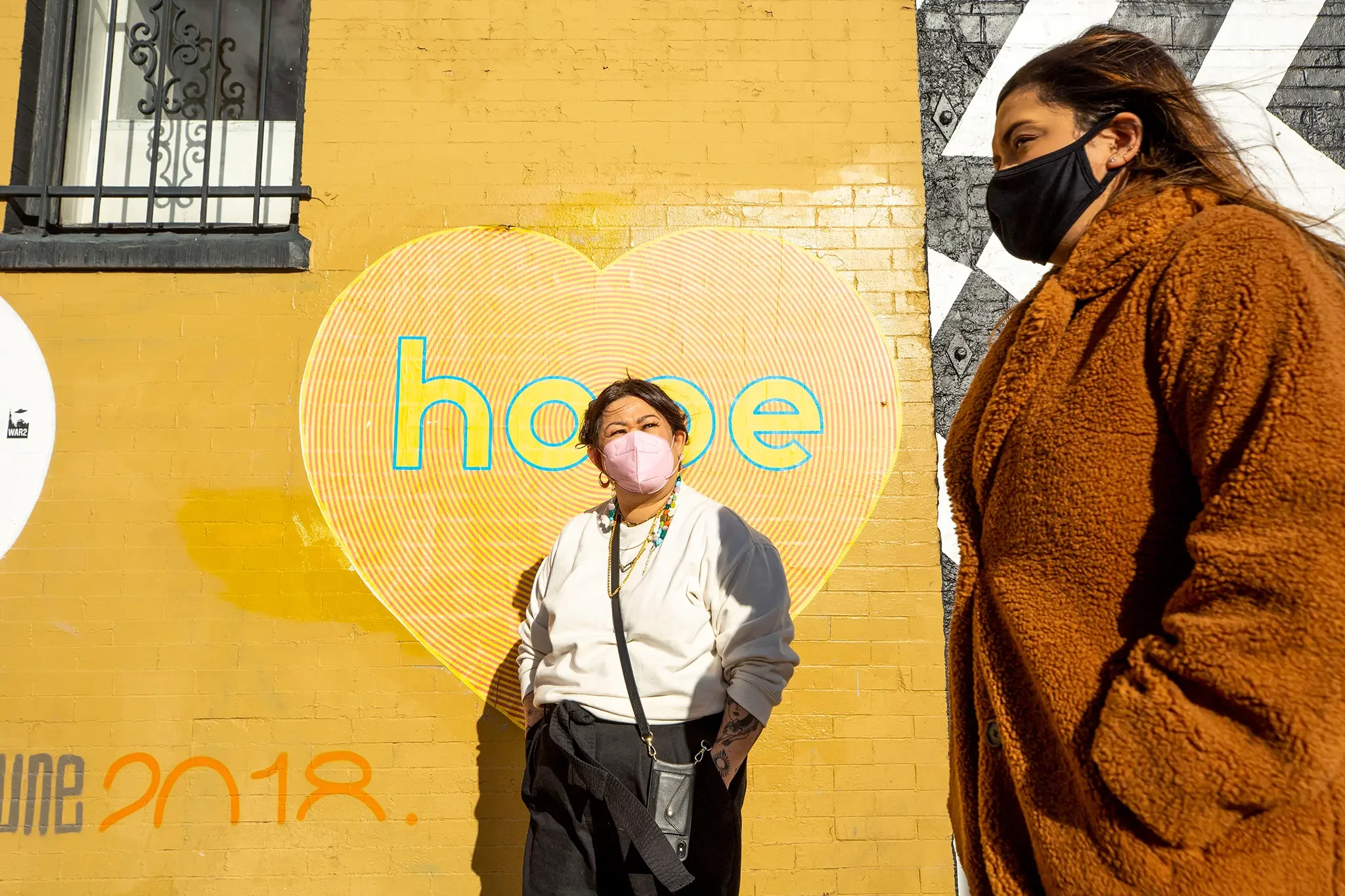 Two women wearing masks stand in front of a yellow brick wall with a large heart-shaped mural that says 'hope' and the date June 2018.