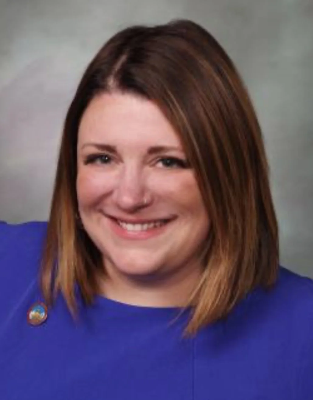 A woman with shoulder-length reddish-brown hair, wearing a blue top, smiling at the camera against a neutral background.