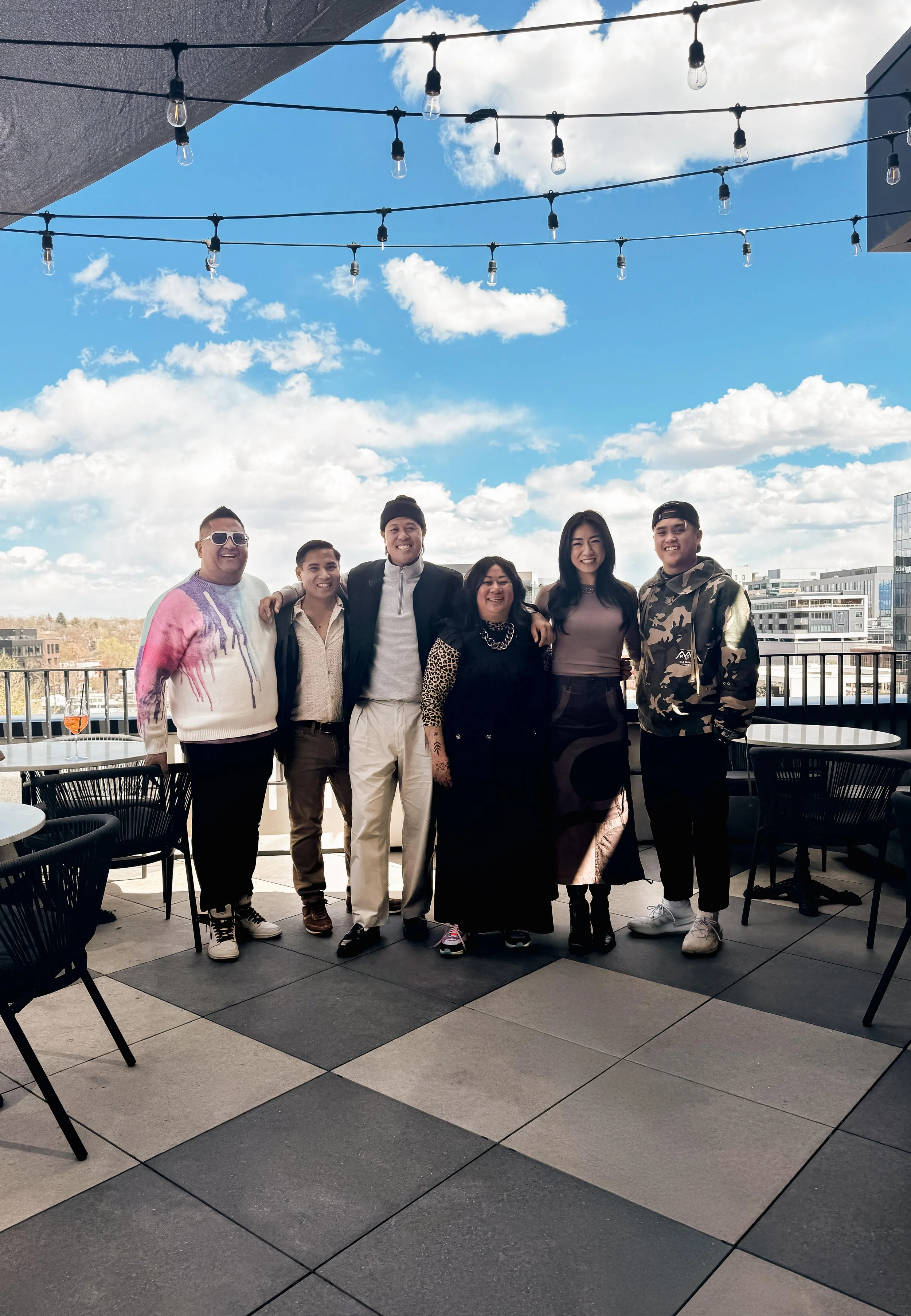 A group of seven diverse people standing on a rooftop terrace with tables and chairs, under a string of outdoor lights, smiling at the camera with a cityscape and partly cloudy sky in the background.