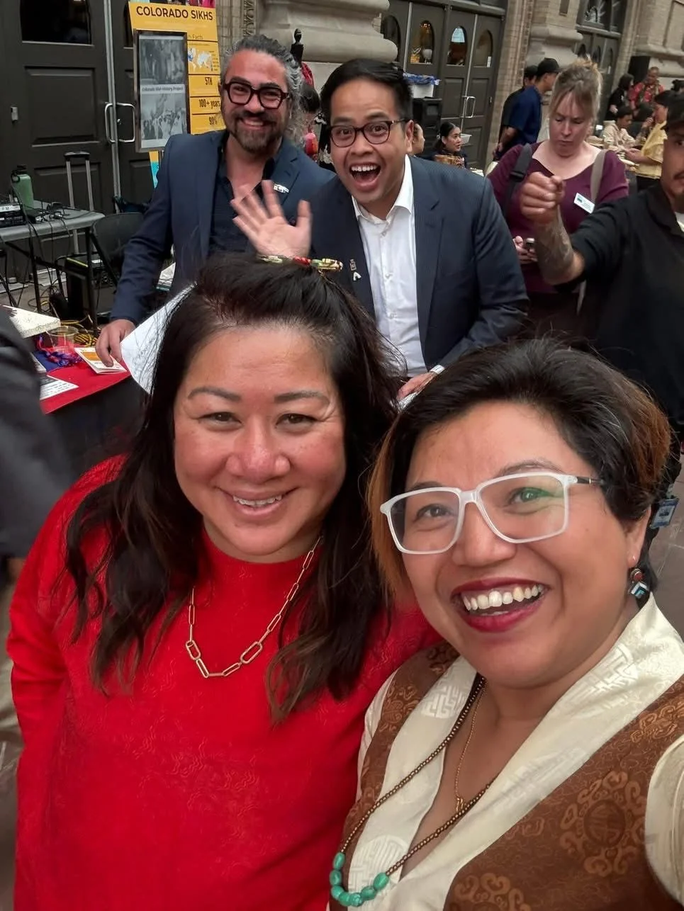 Group of people at an event, smiling for a selfie. Two women are front and center, one in a red top and the other in glasses and a traditional outfit. In the background, two men are smiling and waving, with a colorful sign about Colorado Sikhs visibl