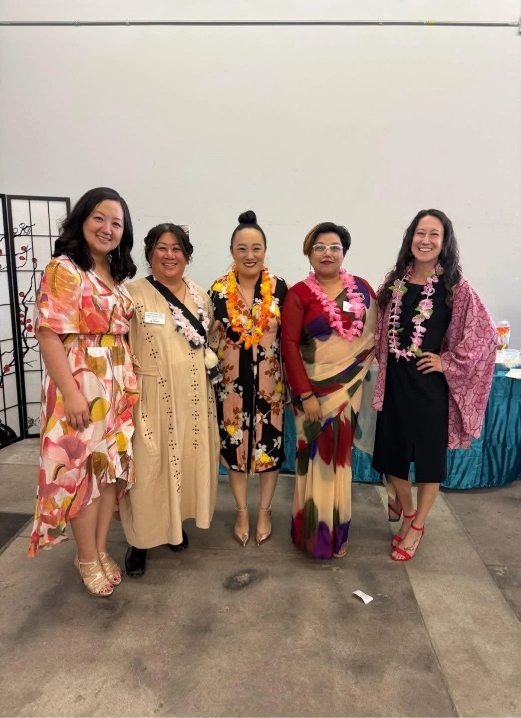 Five women dressed in colorful, floral or festive attire, standing together indoors against a plain white wall, smiling for the camera.