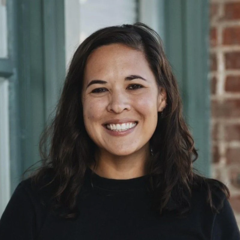 A woman with long dark hair smiling outdoors, wearing a black top, with a building and a window in the background.