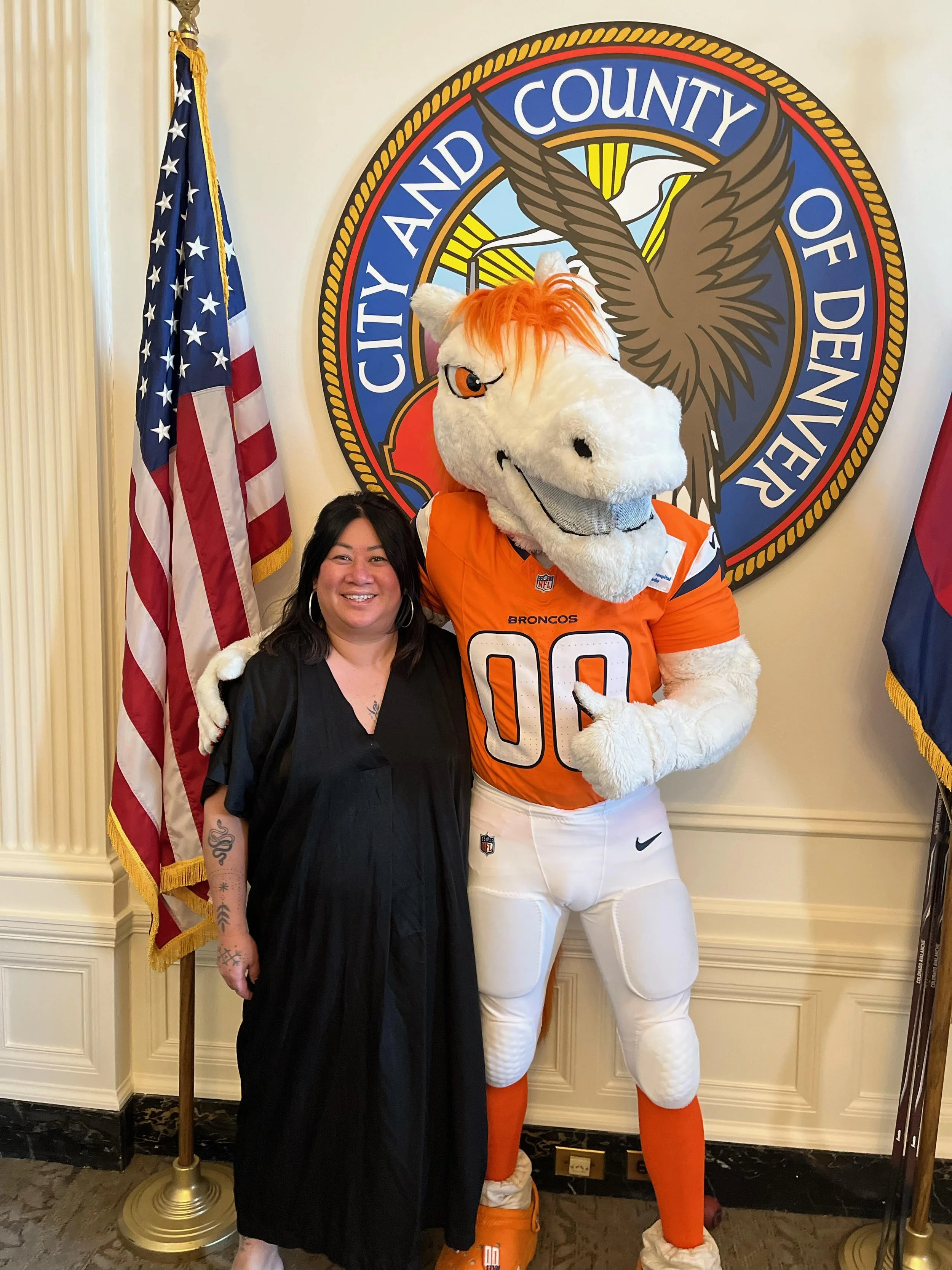 A woman standing next to the Denver Broncos mascot, a white horse with orange hair, in front of a City and County of Denver seal. The woman has dark hair and is wearing a black dress, smiling. The mascot is dressed in NFL football gear with the numbe