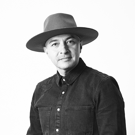 A man wearing a wide-brimmed black hat and a black and white patterned shirt, smiling slightly, in a room with textured light-colored walls and a small mirror.