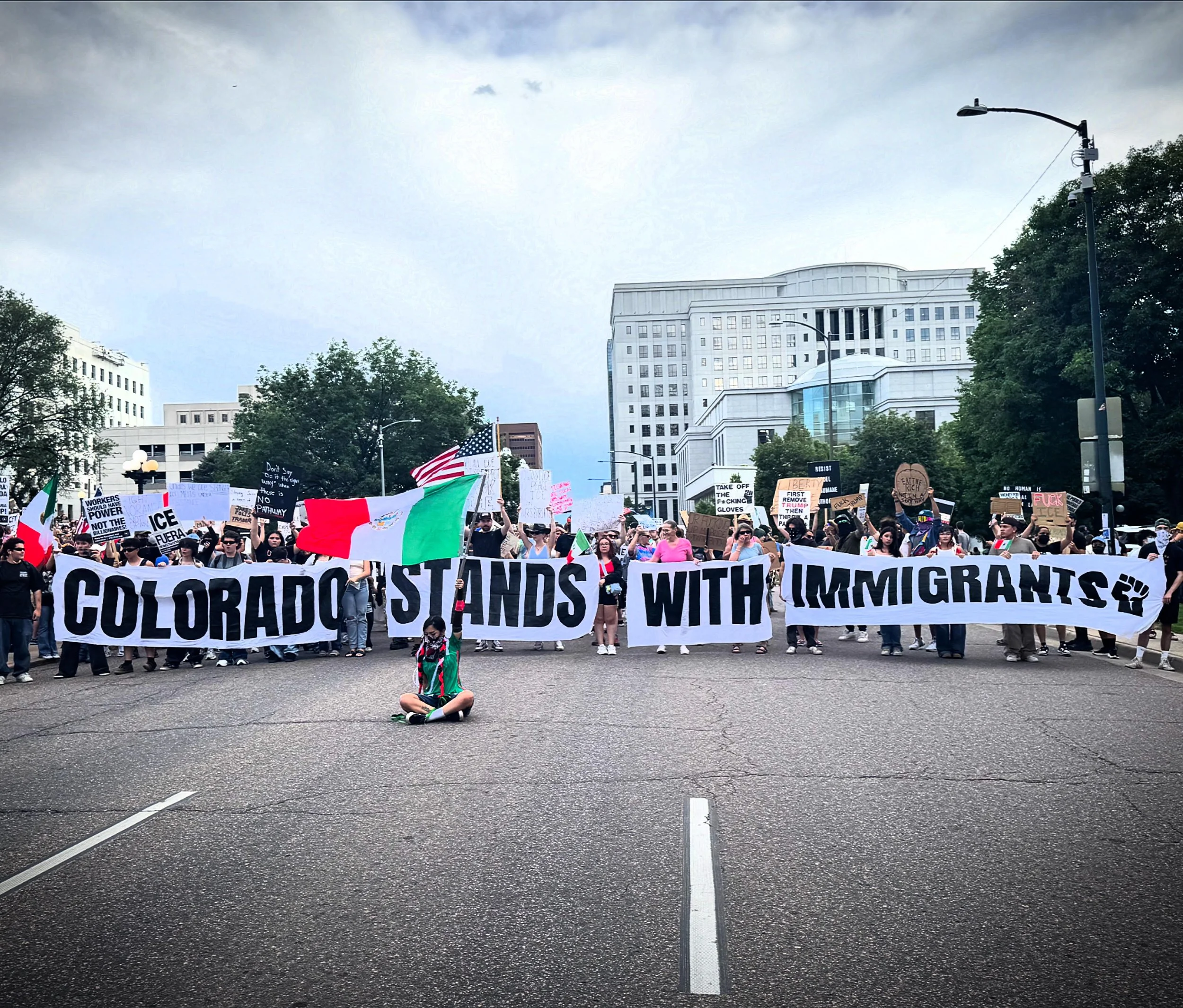 A large group of protesters holding a banner that reads 'Colorado stands with immigrants' on a city street, with many smaller signs and flags.