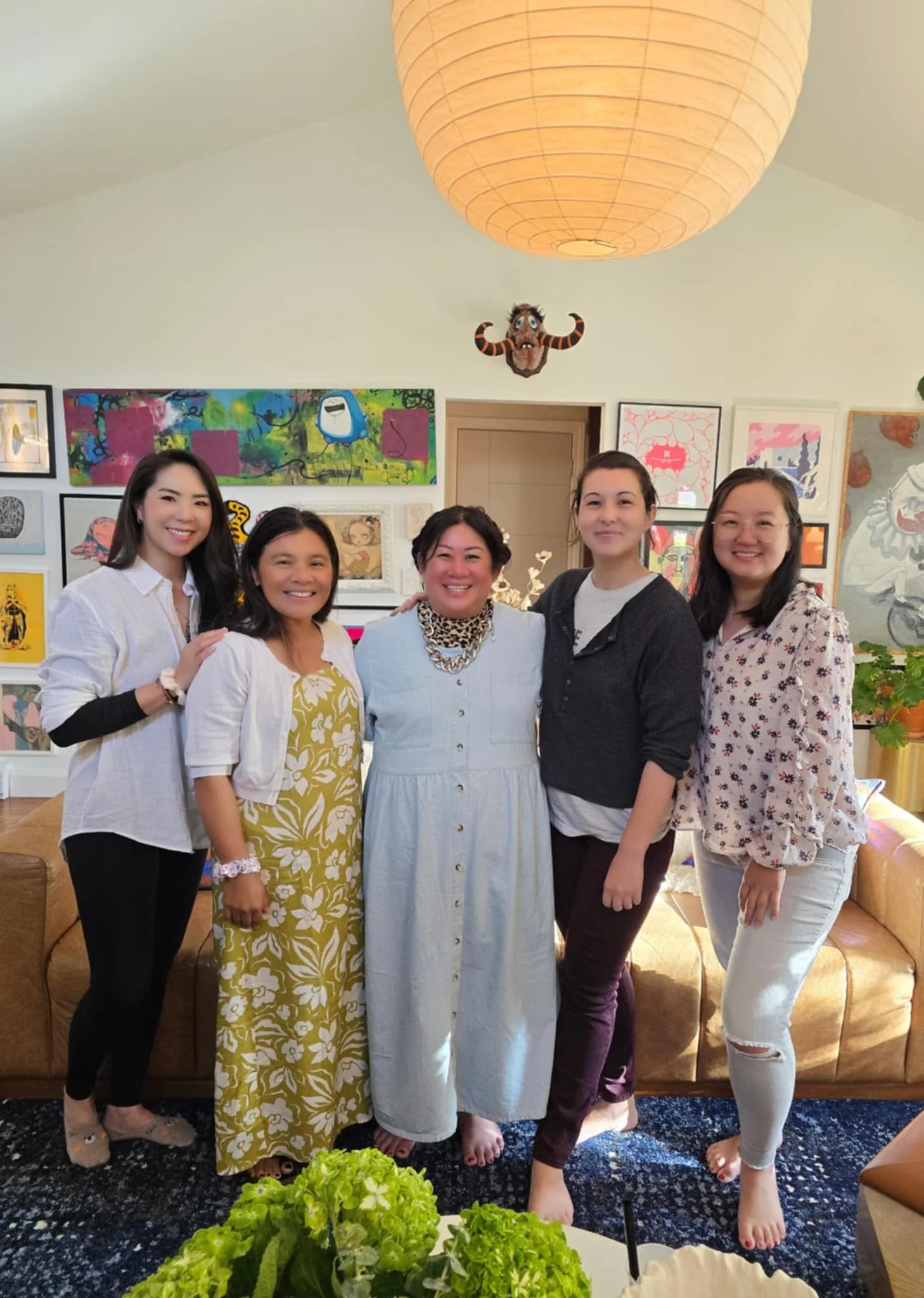 Five women standing together in a cozy living room with artwork on the walls, a tan leather sofa behind them, and a large paper lantern hanging from the ceiling. They are smiling and posing for the camera.