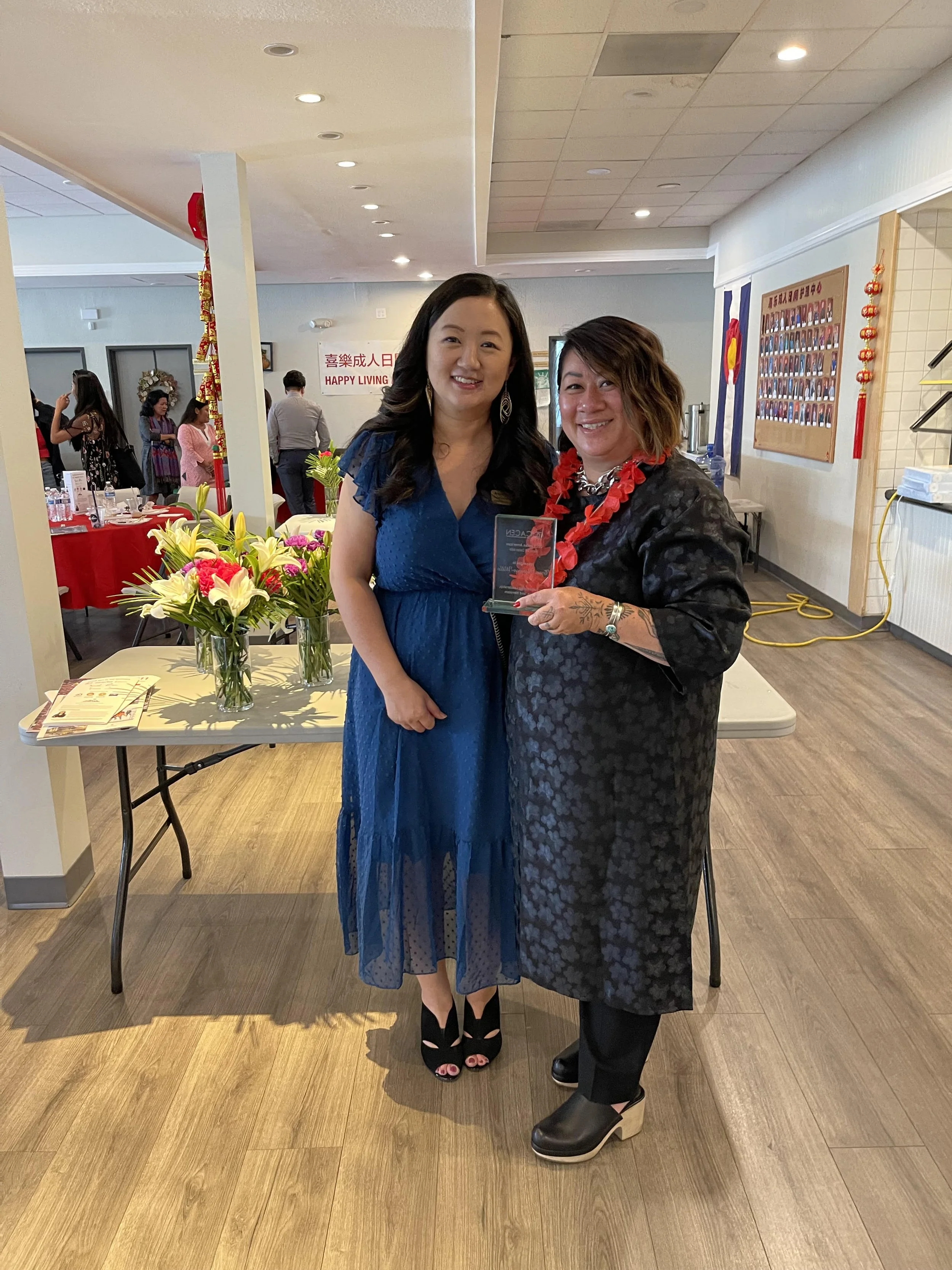 Two women standing together at an indoor event, one holding a glass award, with flowers on a table behind them and other people in the background.