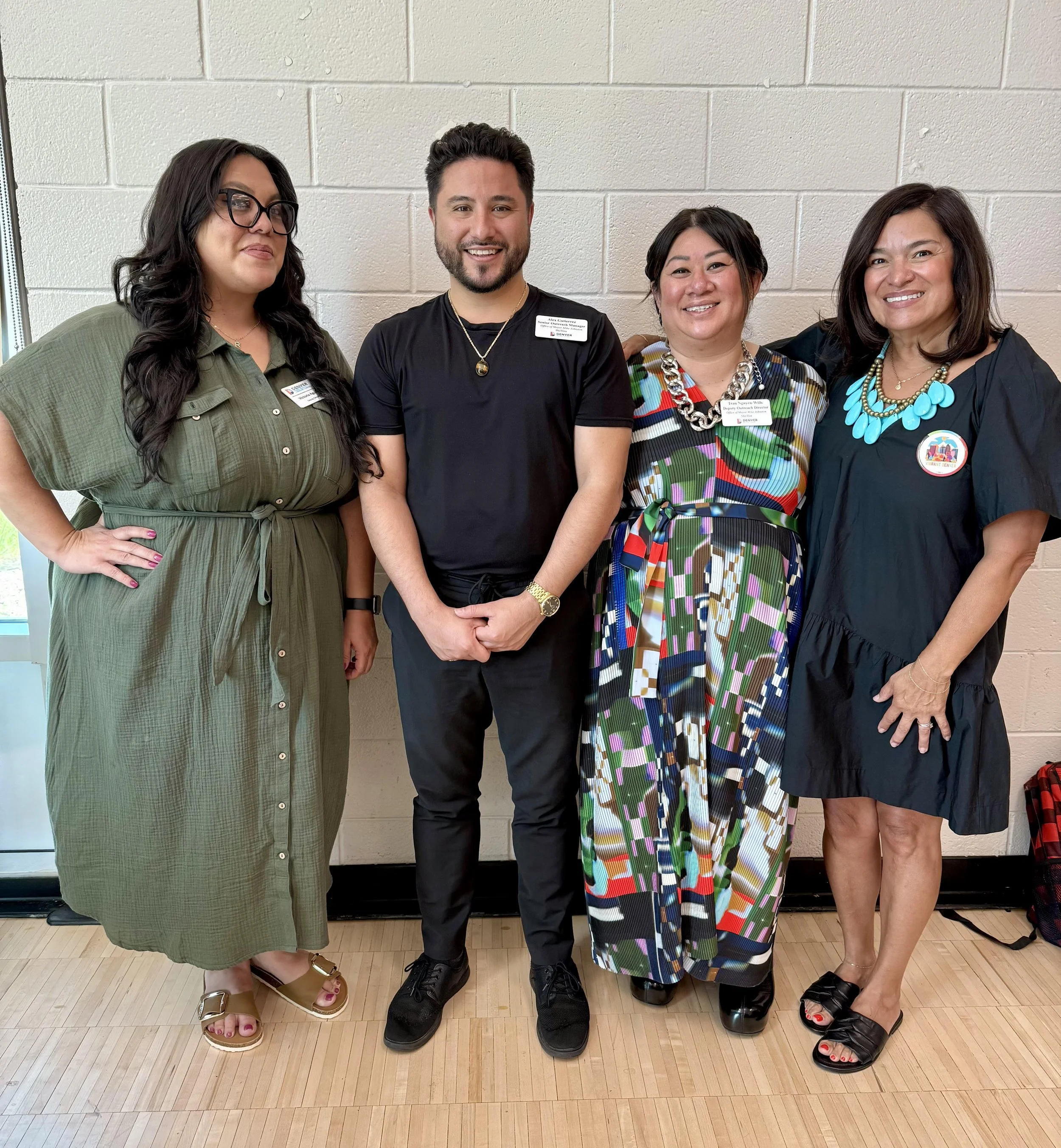 Four people standing side by side indoors, smiling for a photo, against a beige brick wall, dressed in semi-formal and colorful attire.