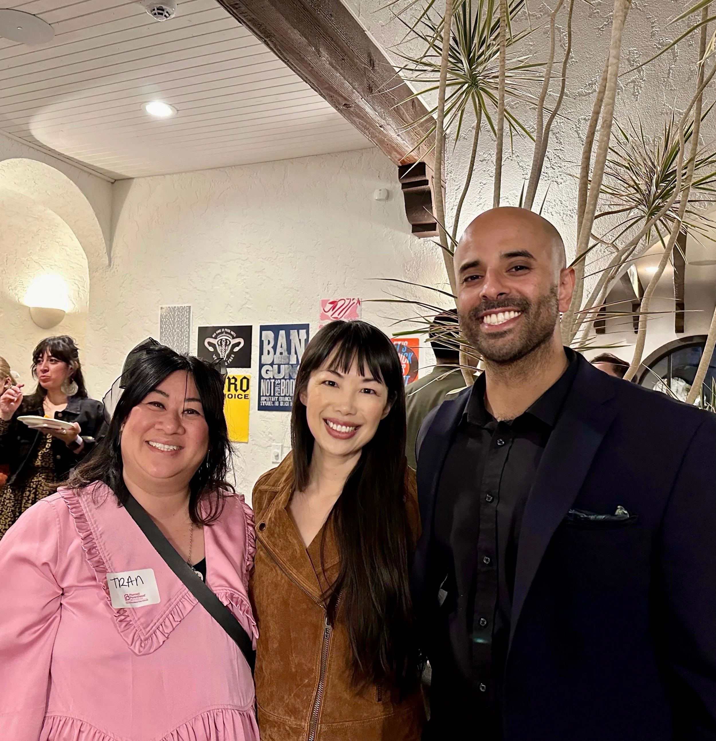 Three people smiling at a social gathering, standing in front of a wall decorated with promotional posters, with a woman in pink, another in brown, and a man in a black suit, indoors with a cozy ambiance.