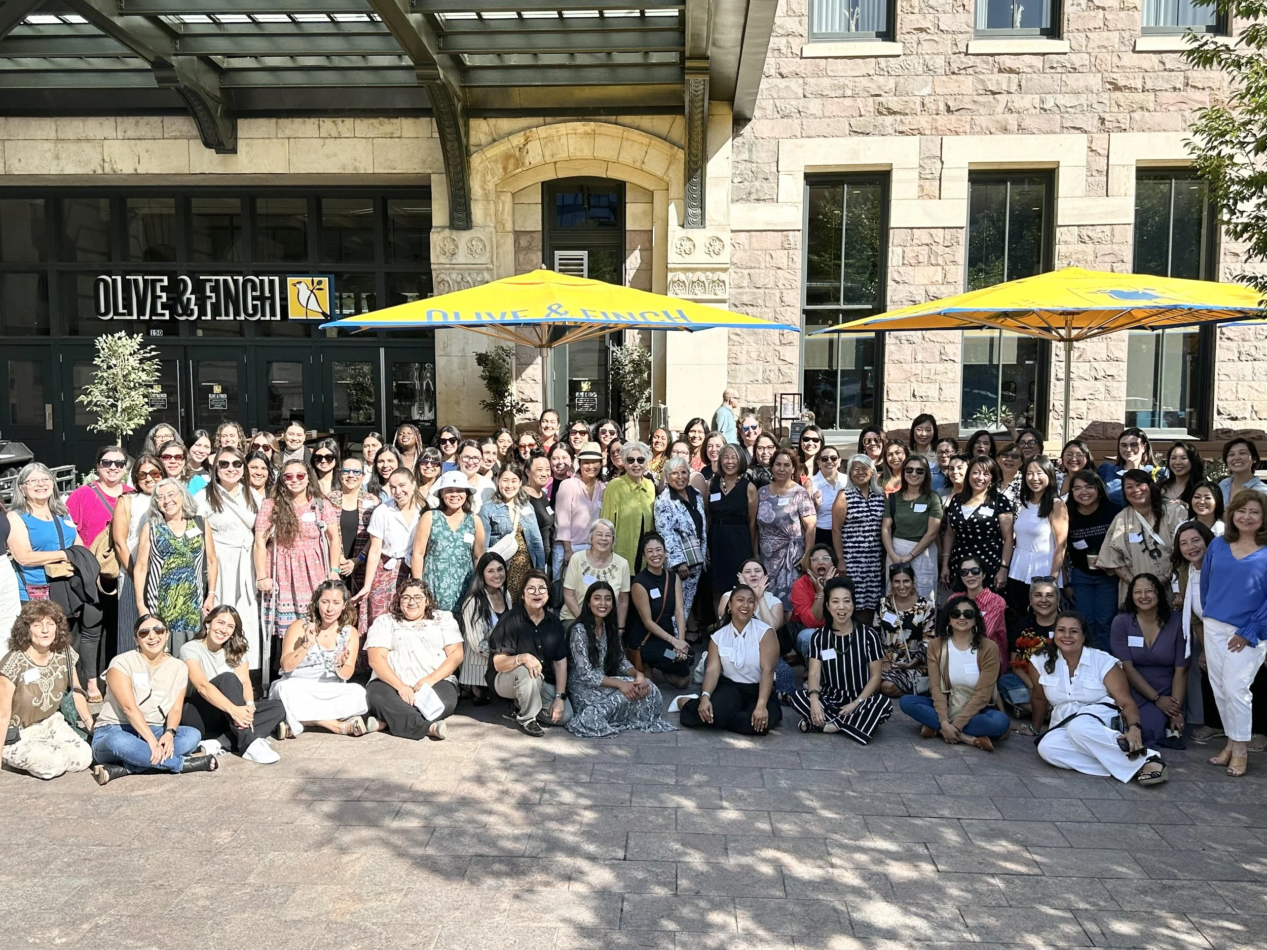 Group of women gathered outside a restaurant called Olive & Finch, some sitting and some standing, with large yellow umbrellas overhead on a sunny day.