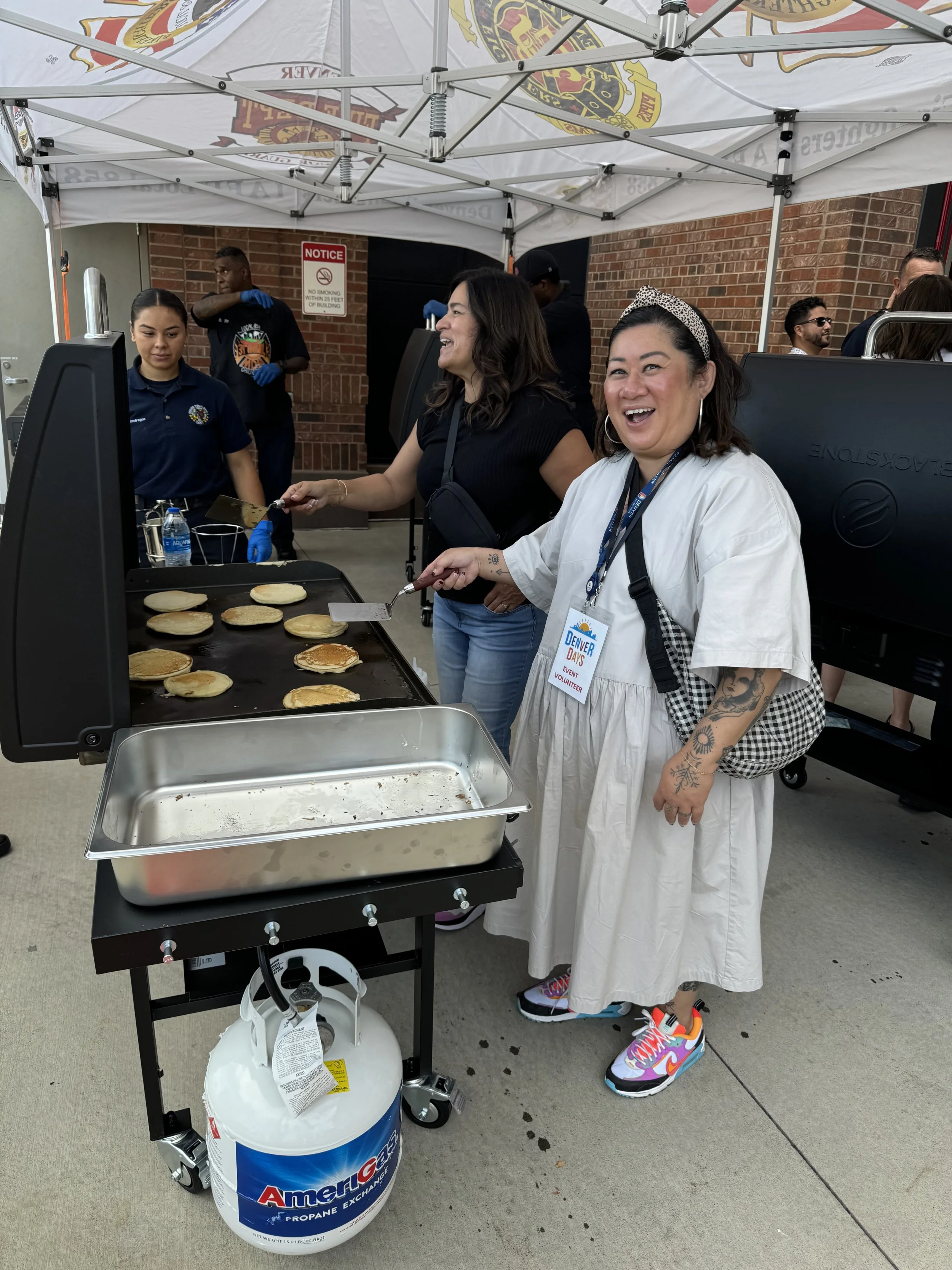 Two women cooking pancakes on a griddle at an outdoor event, with one woman smiling and wearing colorful sneakers. Several people are in the background under a white canopy, and there is a propane tank attached to the grill.
