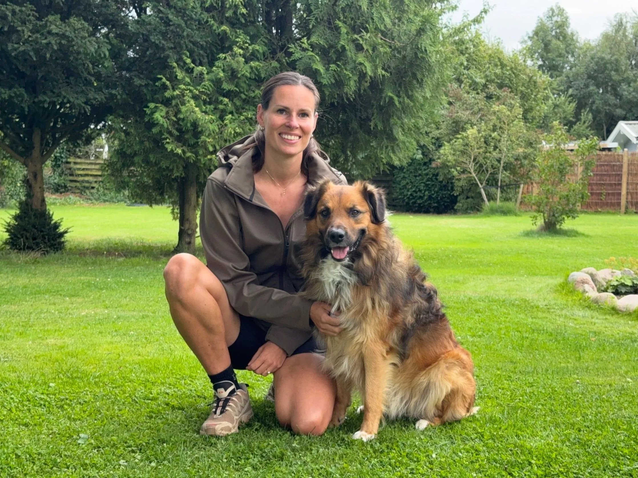 En kvinde og en hund i en grøn have med træer og en farvet træhegnet baggrund.