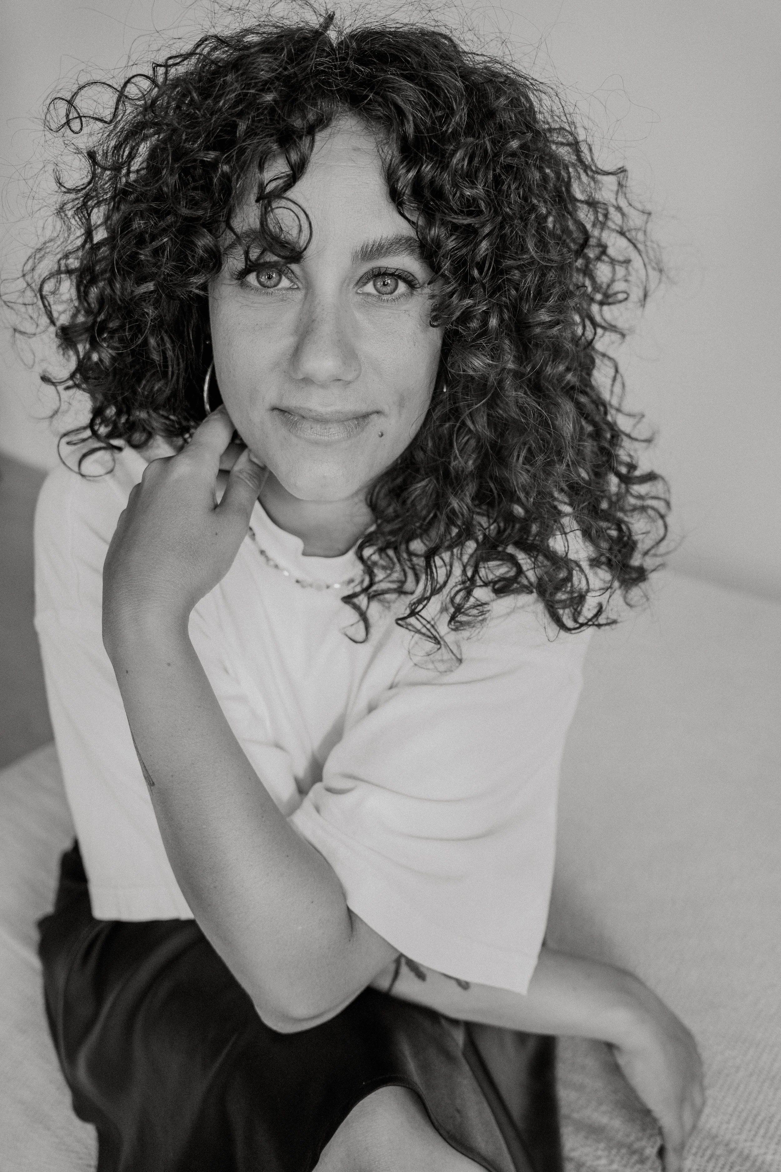 Schwarz-weiße Porträtaufnahme einer Frau mit lockigem Haar, die direkt in die Kamera schaut, mit einer Hand an ihrem Hals. Porträt Shooting im Tageslichtstudio, Mirja Jochum, Fotostudio Saarland., Giulia Posteraro, Branding Fotografie Saarland.
